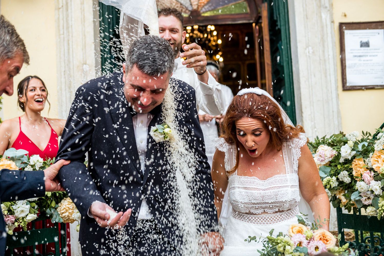 Wedding guests throw rice and confetti outside a cream-colored church entrance.