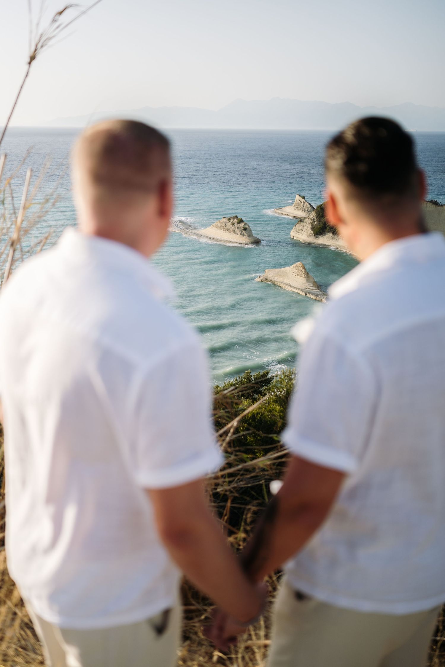 Two people in white shirts overlooking dramatic coastal rock formations and blue ocean waters.