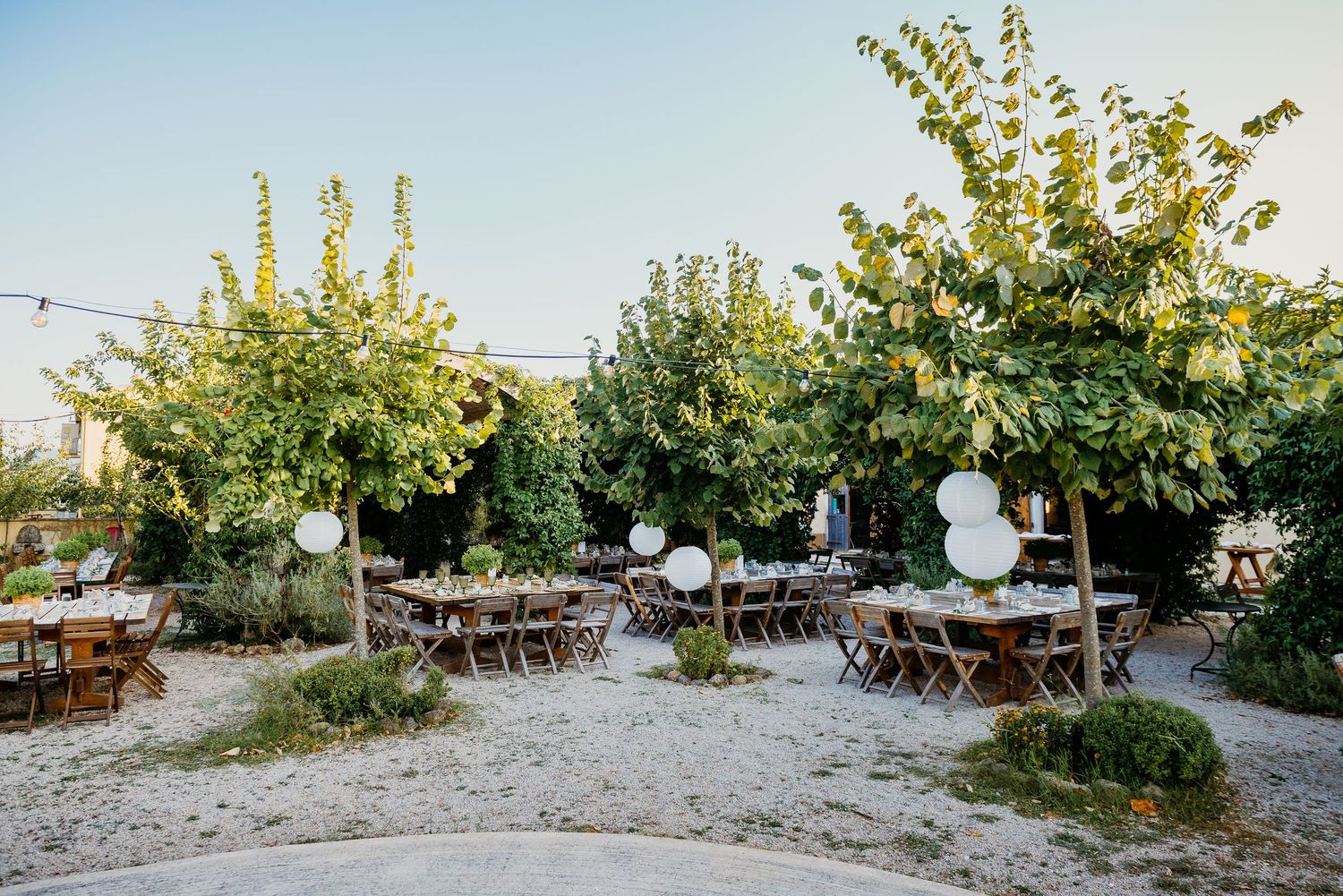 Long wooden tables arranged outdoors for a wedding reception with natural garden surroundings.