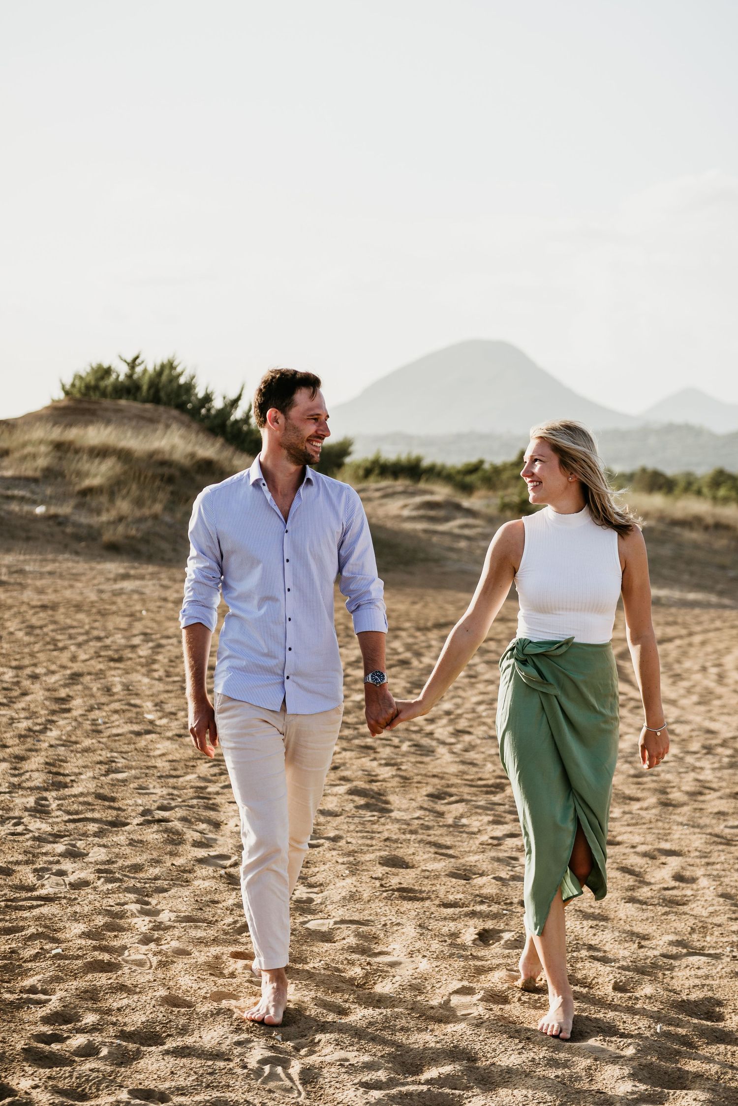 A couple walks hand in hand on a sandy beach with mountains in the background.