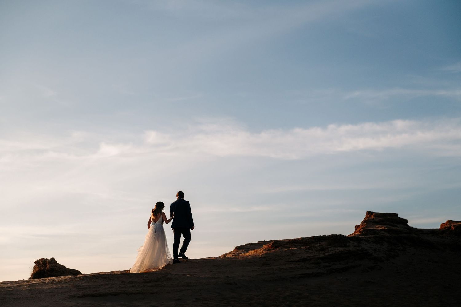 A couple stands silhouetted against a dramatic sunset sky on a rocky hilltop.