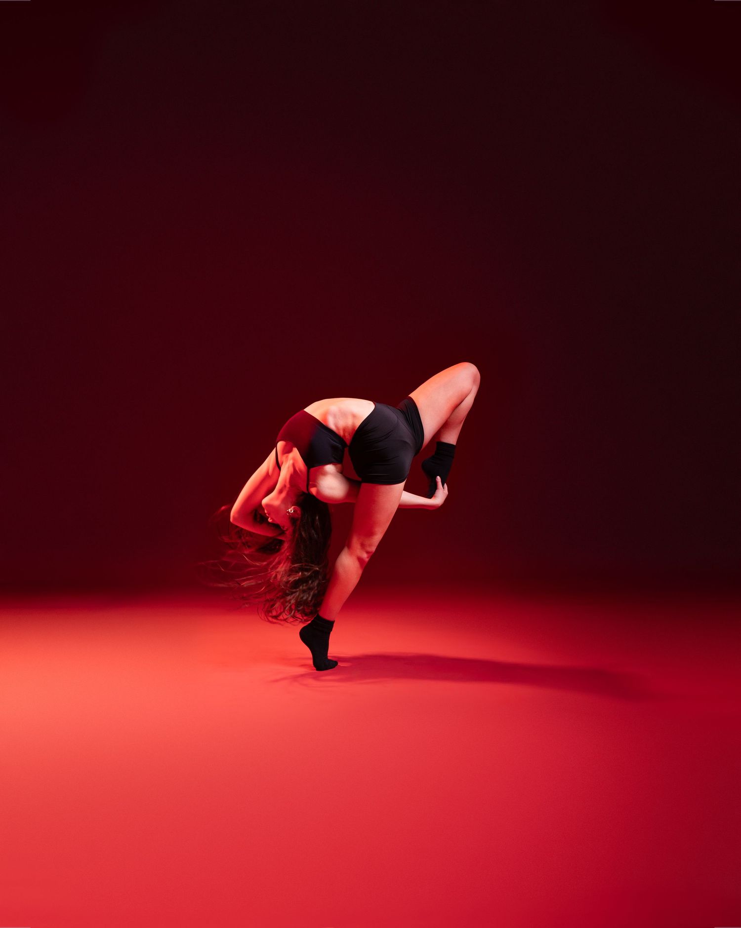 A dancer in black activewear bends dramatically forward on a red-lit studio floor against a dark crimson background.