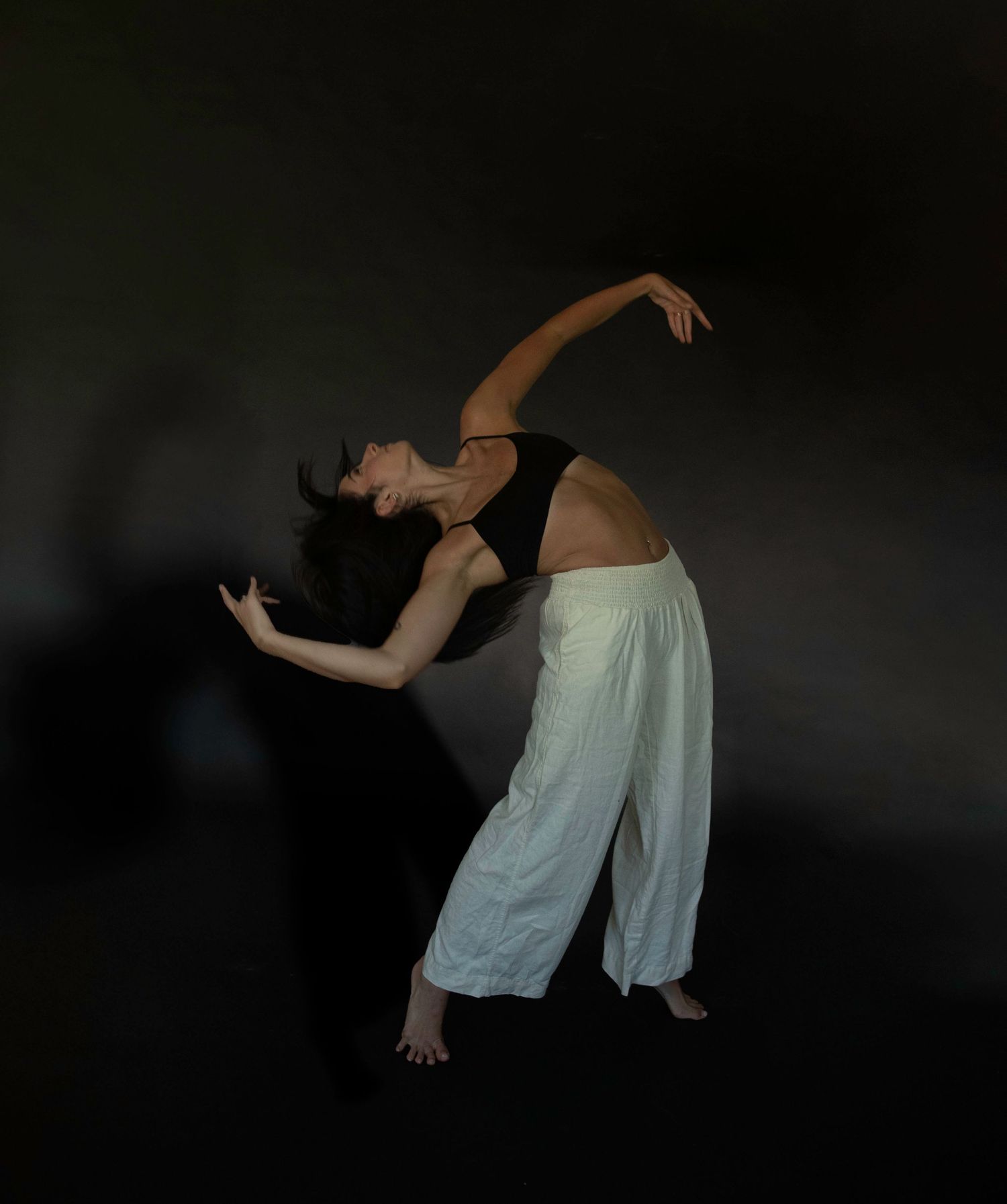 Contemporary dancer in flowing white pants performing expressive movement against dark background.