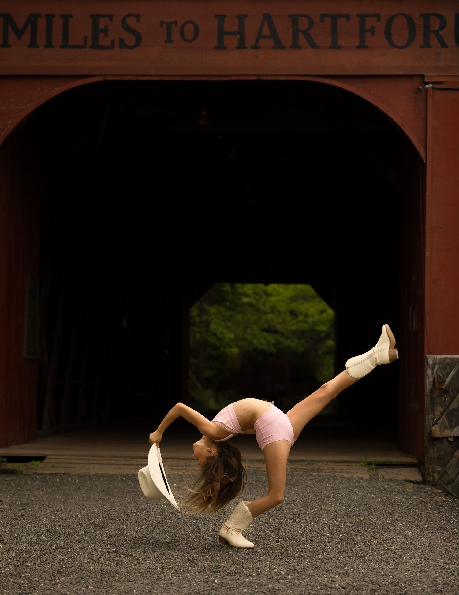 Dancer performing a backbend with leg raised near a red covered bridge with Miles to Hartford sign.