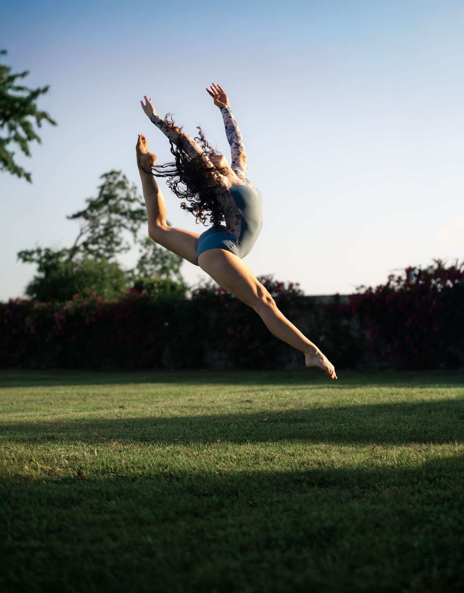 A dancer leaps gracefully in a split jump outdoors on green grass, arms raised, wearing a blue leotard at dusk.