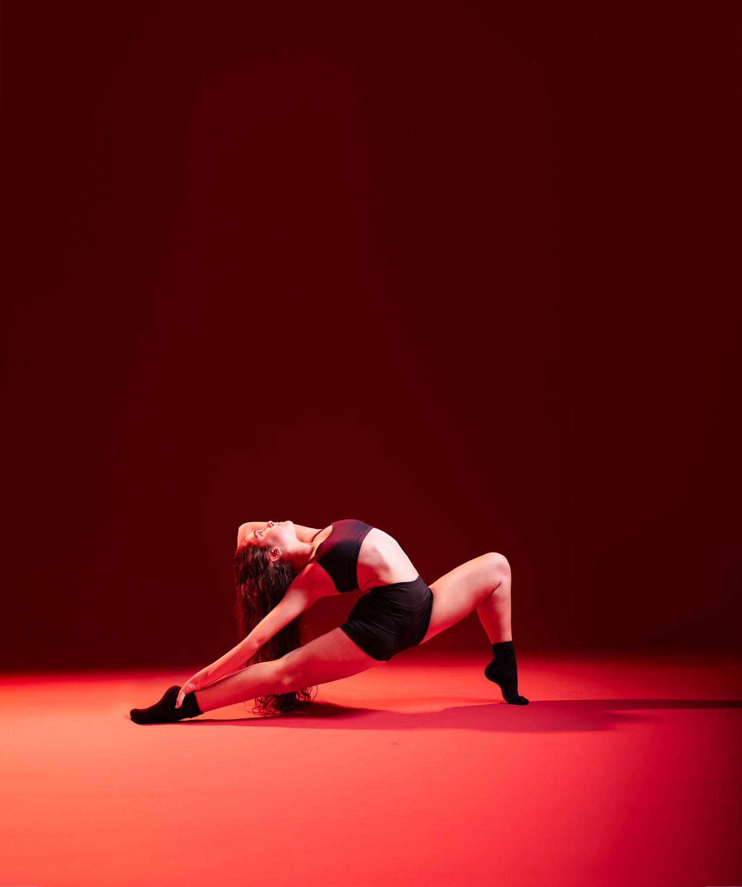 A flexible dancer in black activewear performs a dramatic floor pose against a vibrant red studio background.