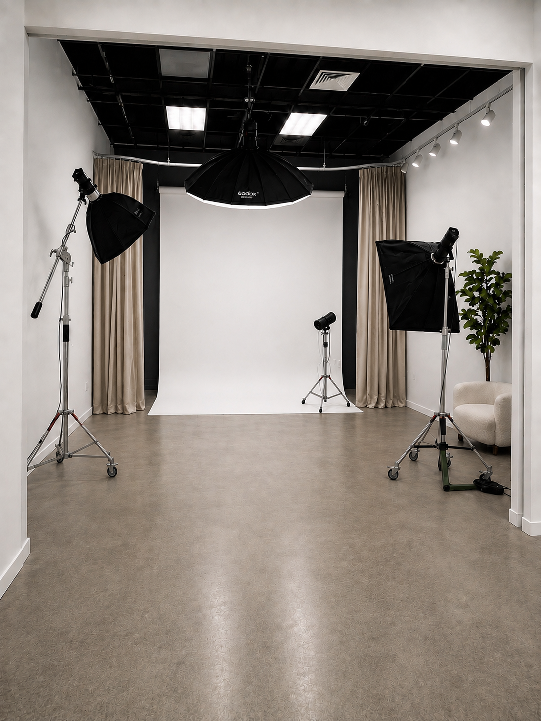 Professional photography studio with white backdrop, studio lights on stands, beige curtains, and a small seating area with a plant.