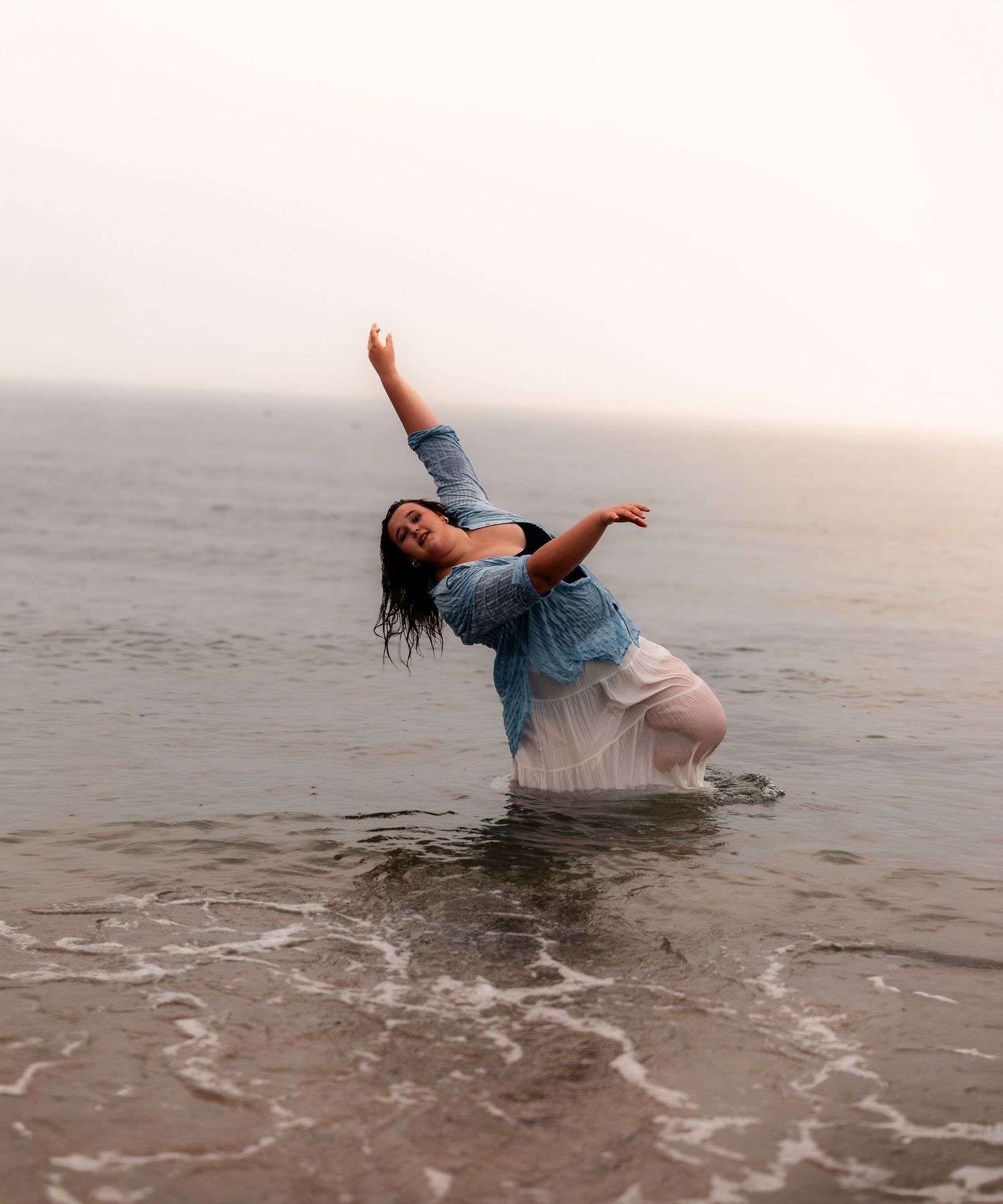 Woman in wet blue shirt dancing gracefully in ocean waves on a misty beach.