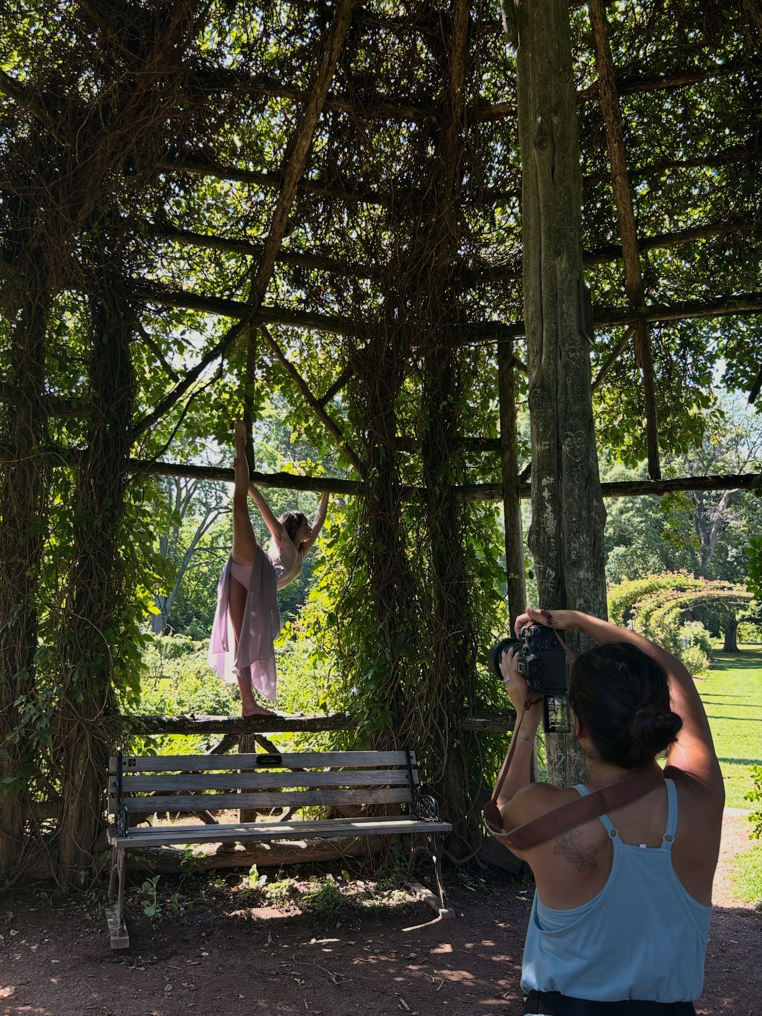 Photographer captures a dancer posing gracefully in a vine-covered pergola beside a white bench in a lush garden.