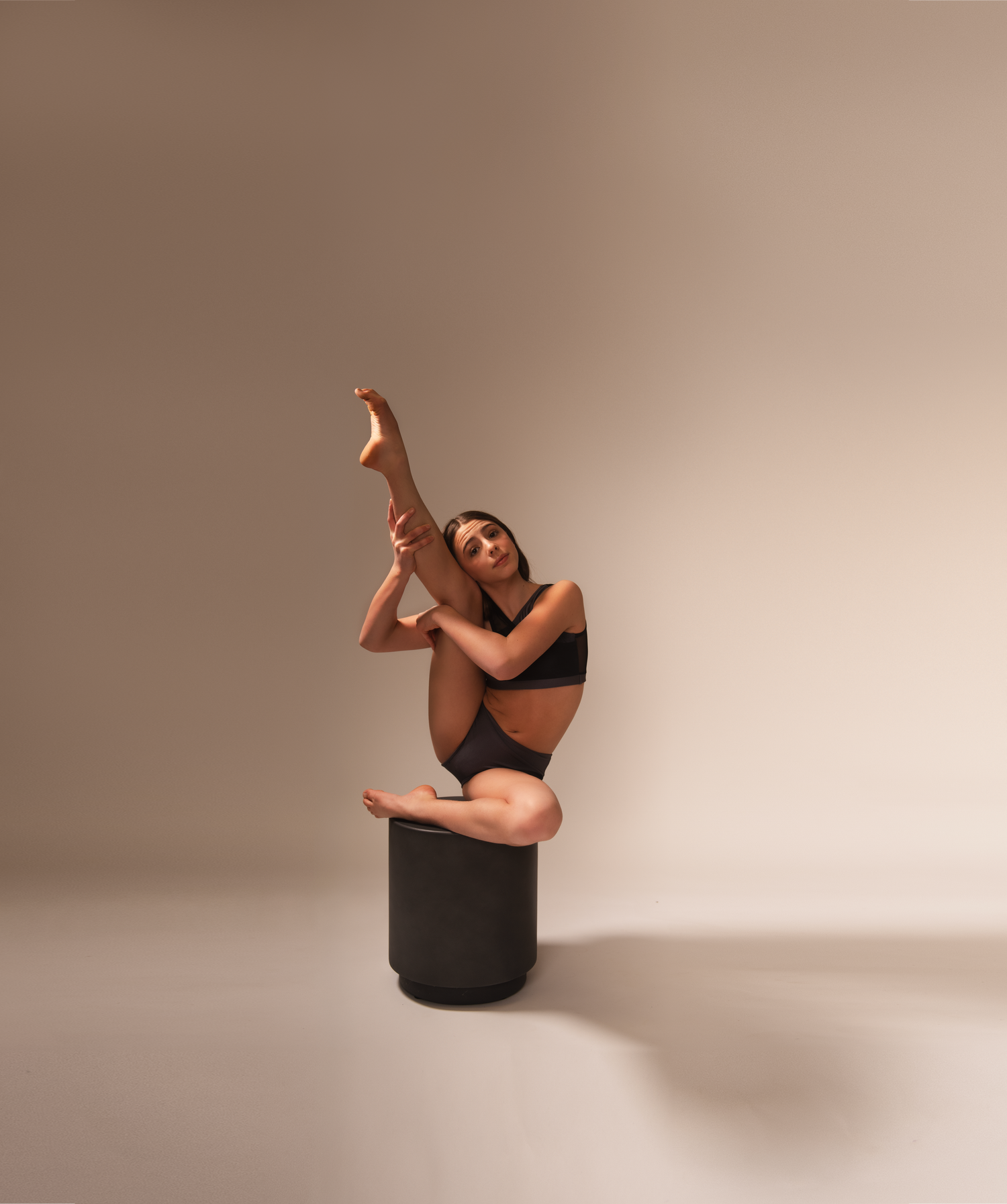 Dancer performs artistic pose while balanced on black cylinder against neutral backdrop.