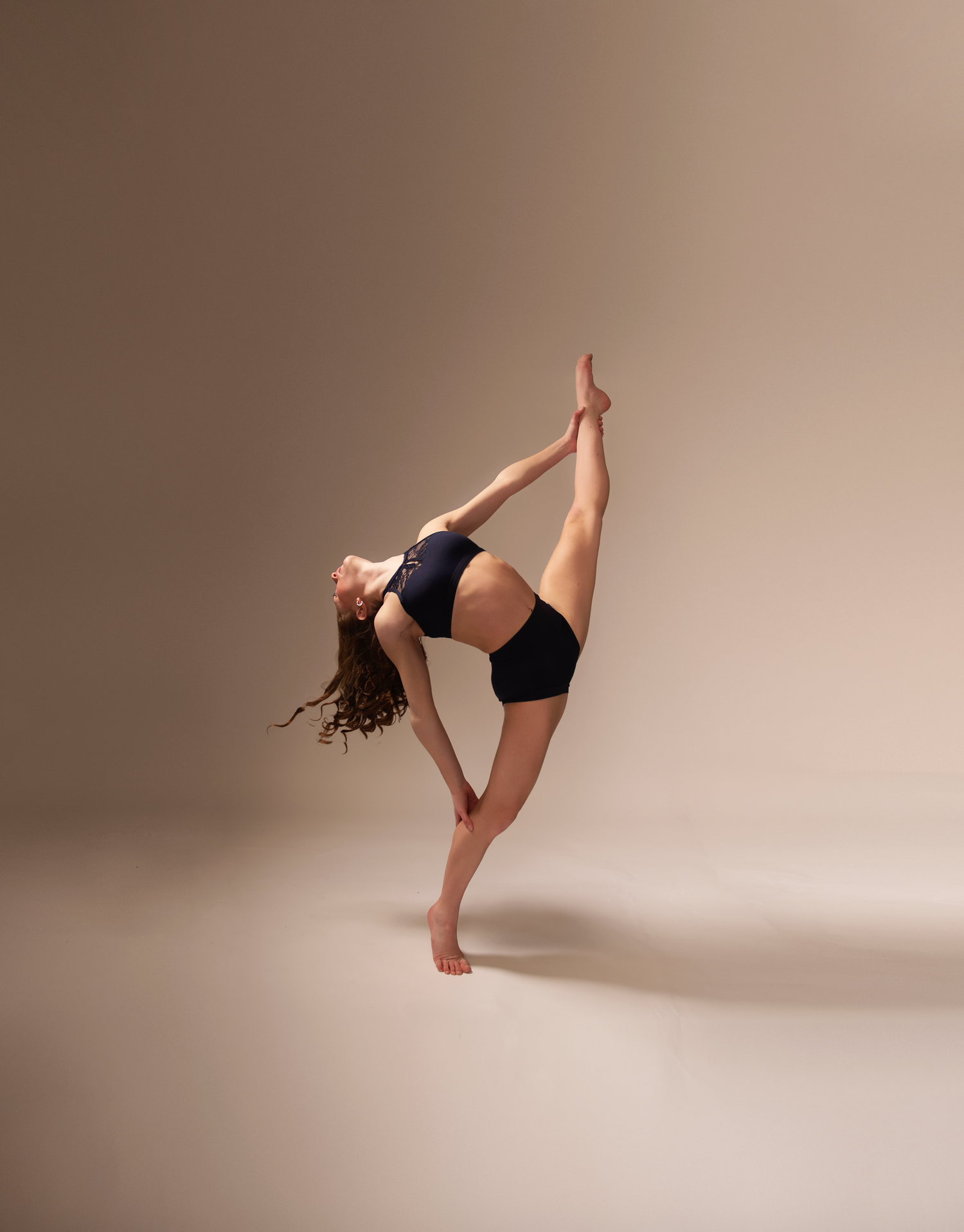 Dancer performs standing split pose in black athletic wear against neutral backdrop.