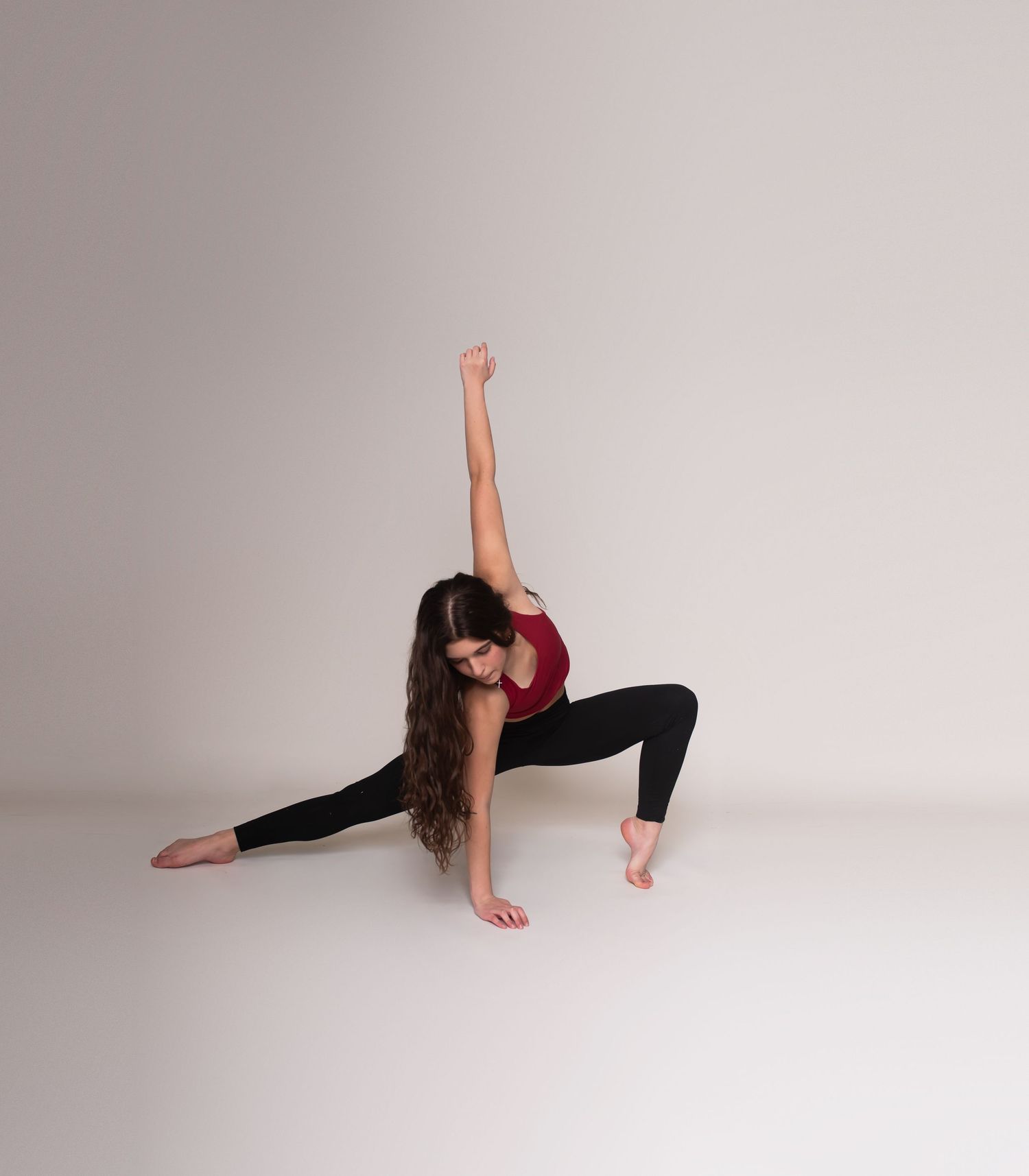 Dancer in red top and black leggings performs a low lunge pose with raised arm against neutral background.
