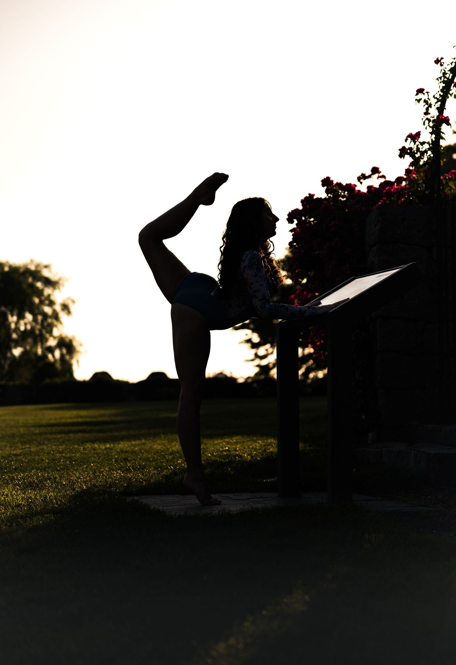 Silhouette of a flexible woman performing a standing split yoga pose outdoors at sunset near flowering plants.