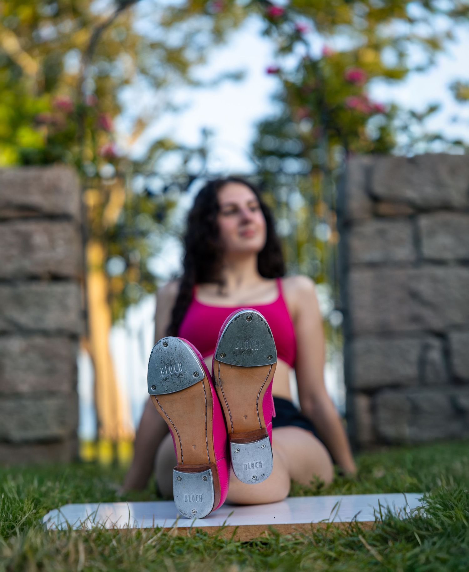 Woman in pink top sitting on grass with tap dance shoes soles facing camera in outdoor park setting.