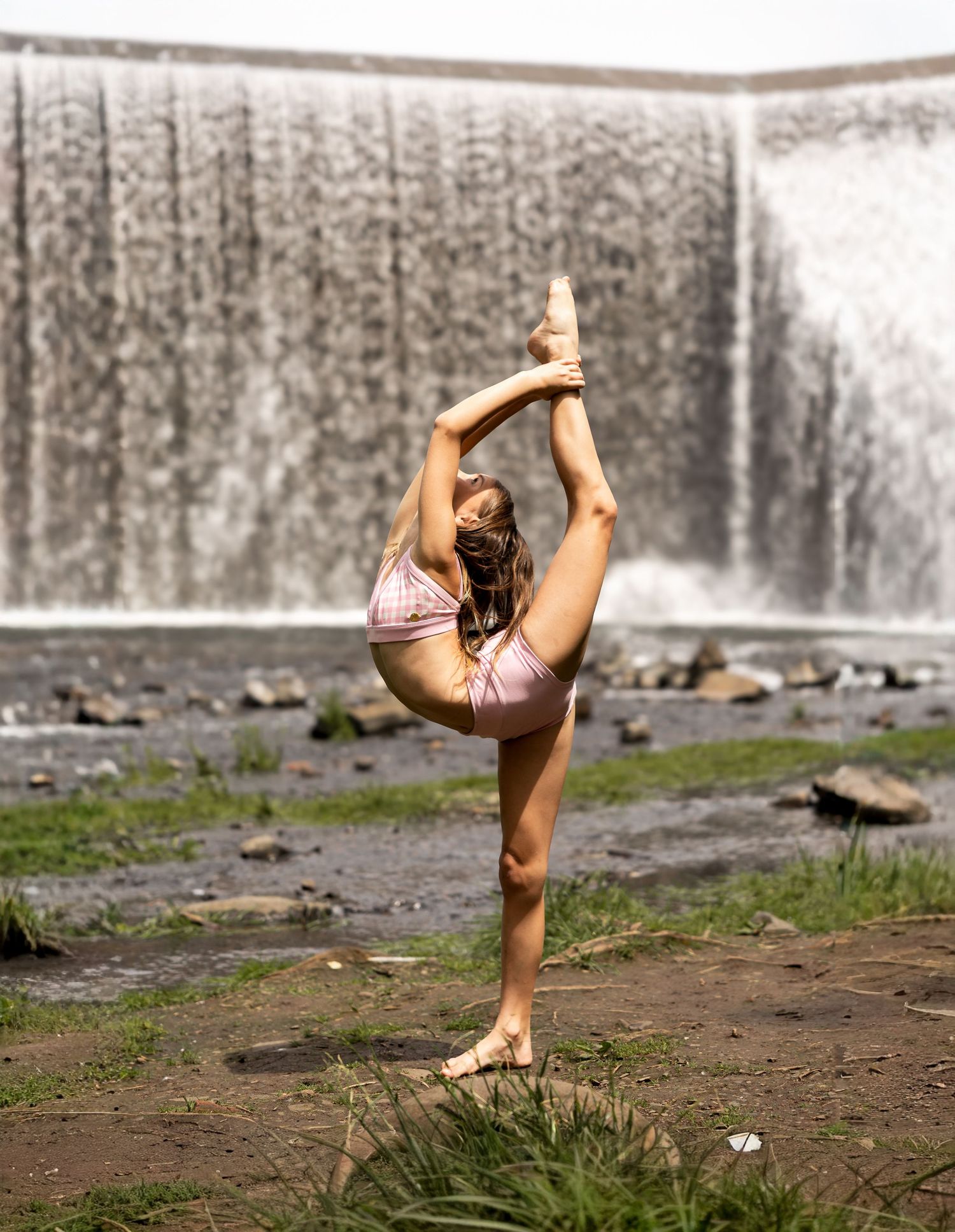 Flexible dancer performs a standing split pose outdoors near a waterfall, wearing a pink leotard.