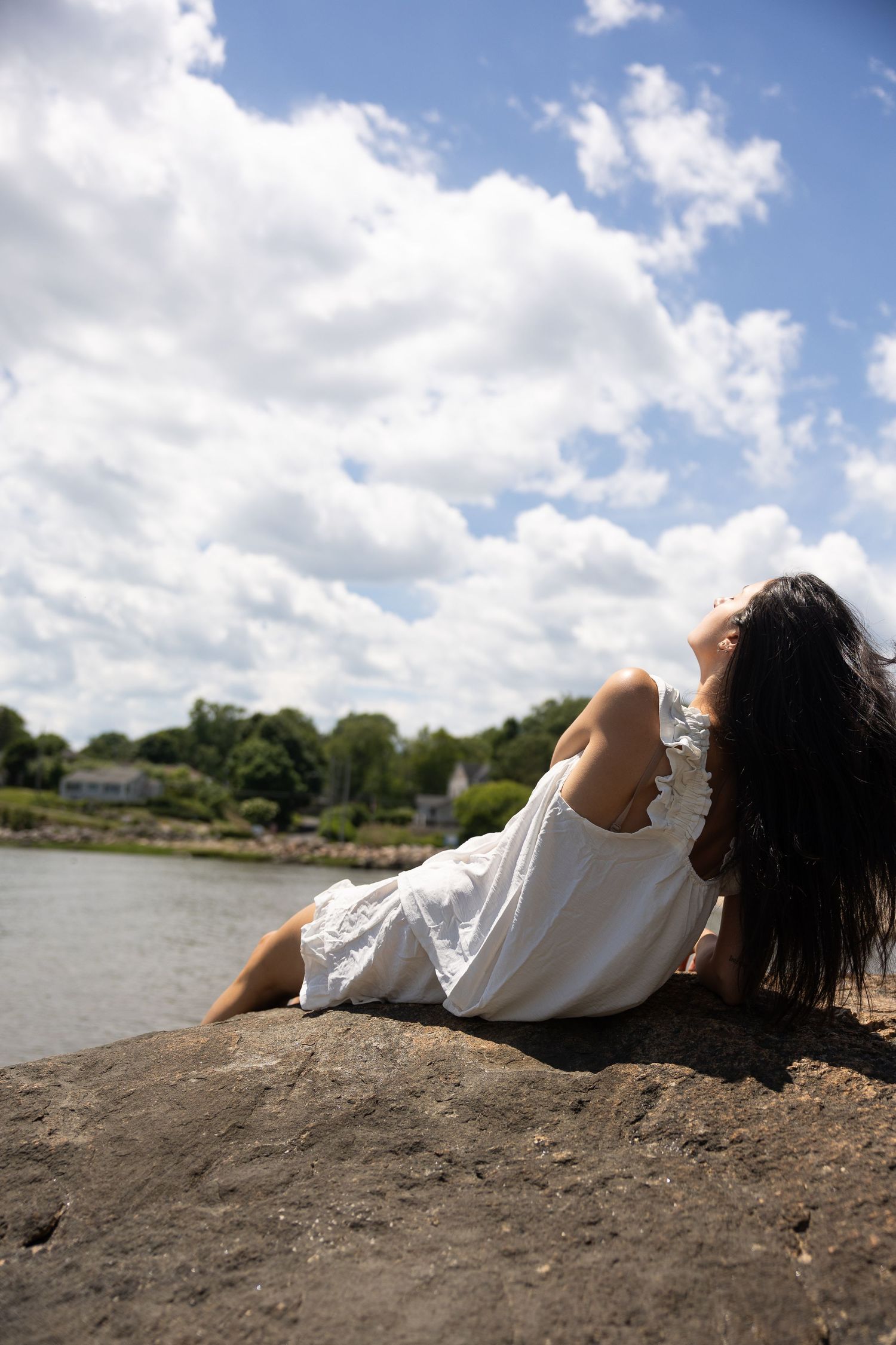 Woman in white dress relaxing on rocks by the water under a cloudy blue sky on a sunny day.