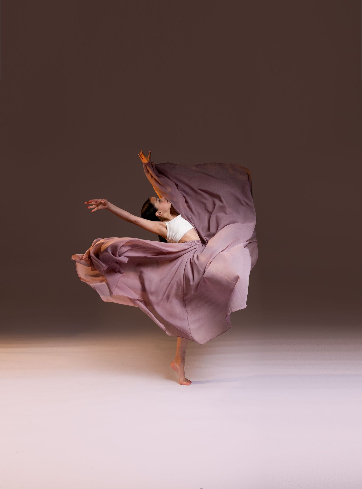 A dancer leaps gracefully in a flowing mauve skirt against a brown studio backdrop.