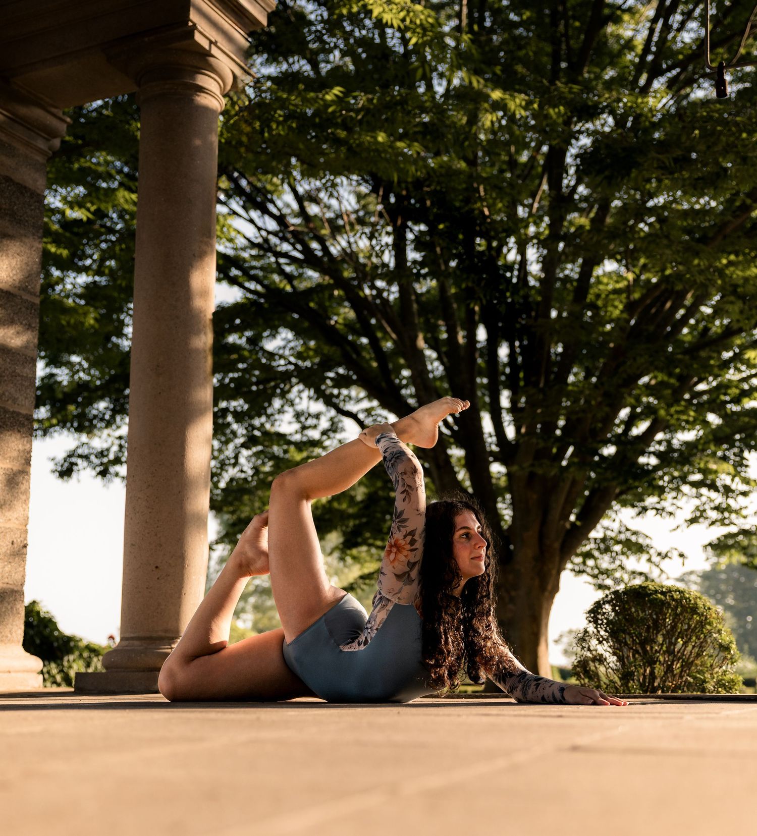 Woman practicing yoga in a flexible pose under stone columns surrounded by lush green trees outdoors.