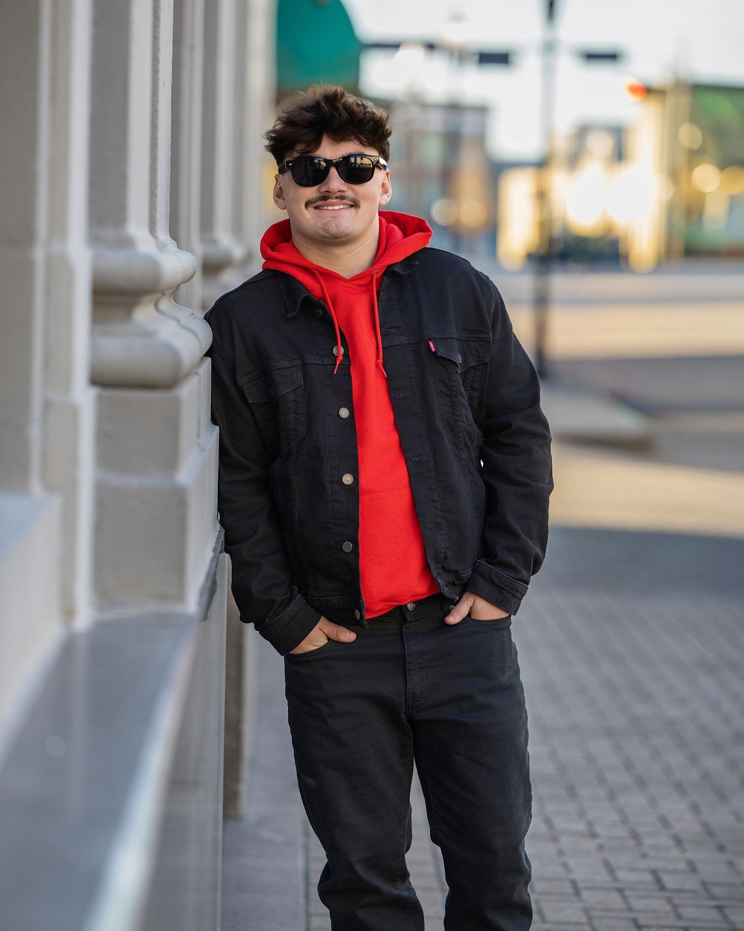 A person in a black jacket and red hoodie poses confidently against a modern architectural backdrop in an urban setting.