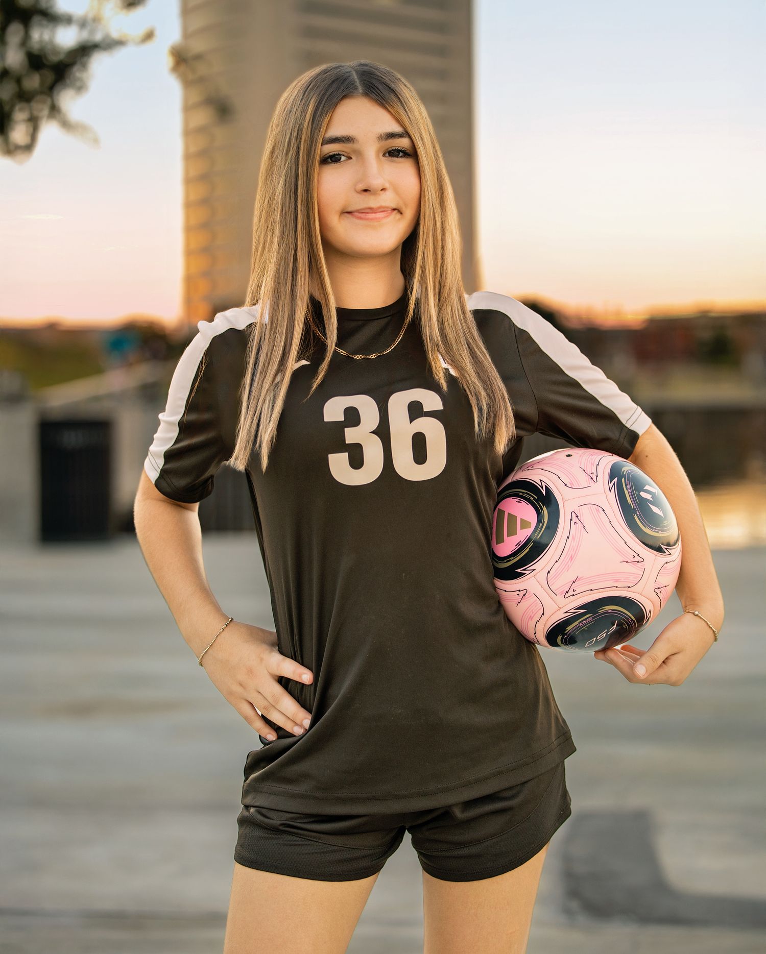 Soccer player wearing black jersey number 36 poses with pink ball during golden hour sunset.