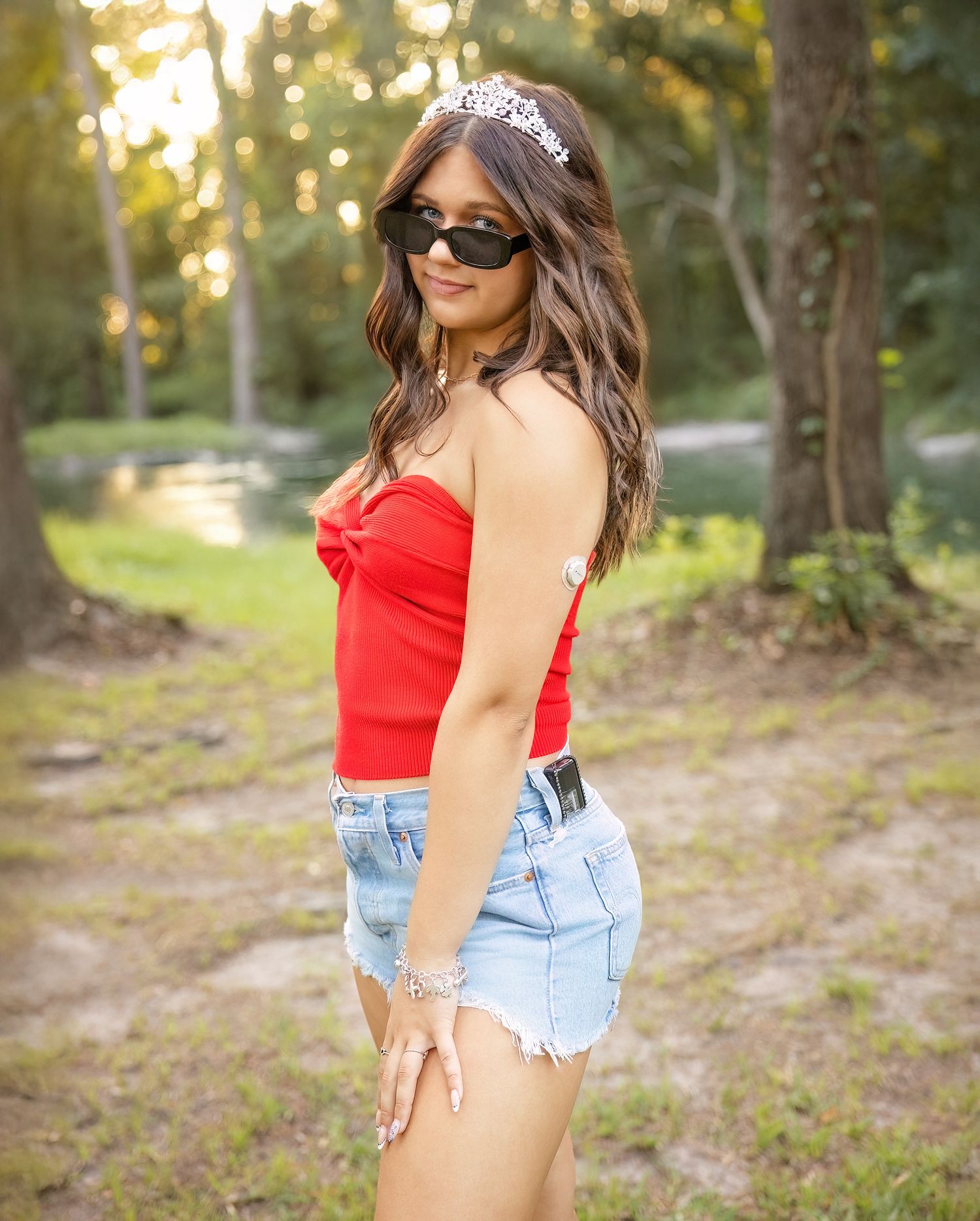 A woman in a red tank top and denim shorts poses casually in a sunlit wooded area during golden hour.