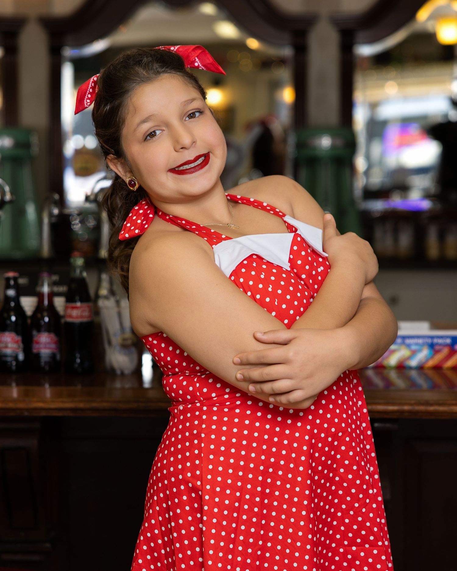 A person in a red and white polka dot dress poses playfully against a dark restaurant interior background.