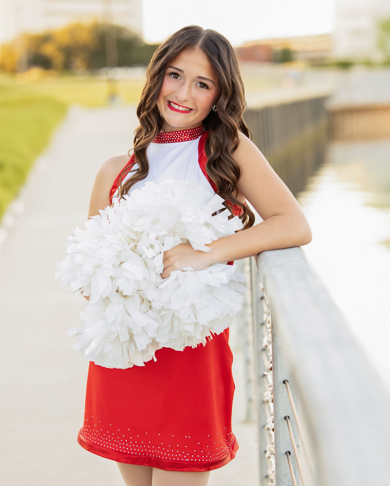 Cheerleader in red and white uniform performs with white pom poms outdoors near railing.