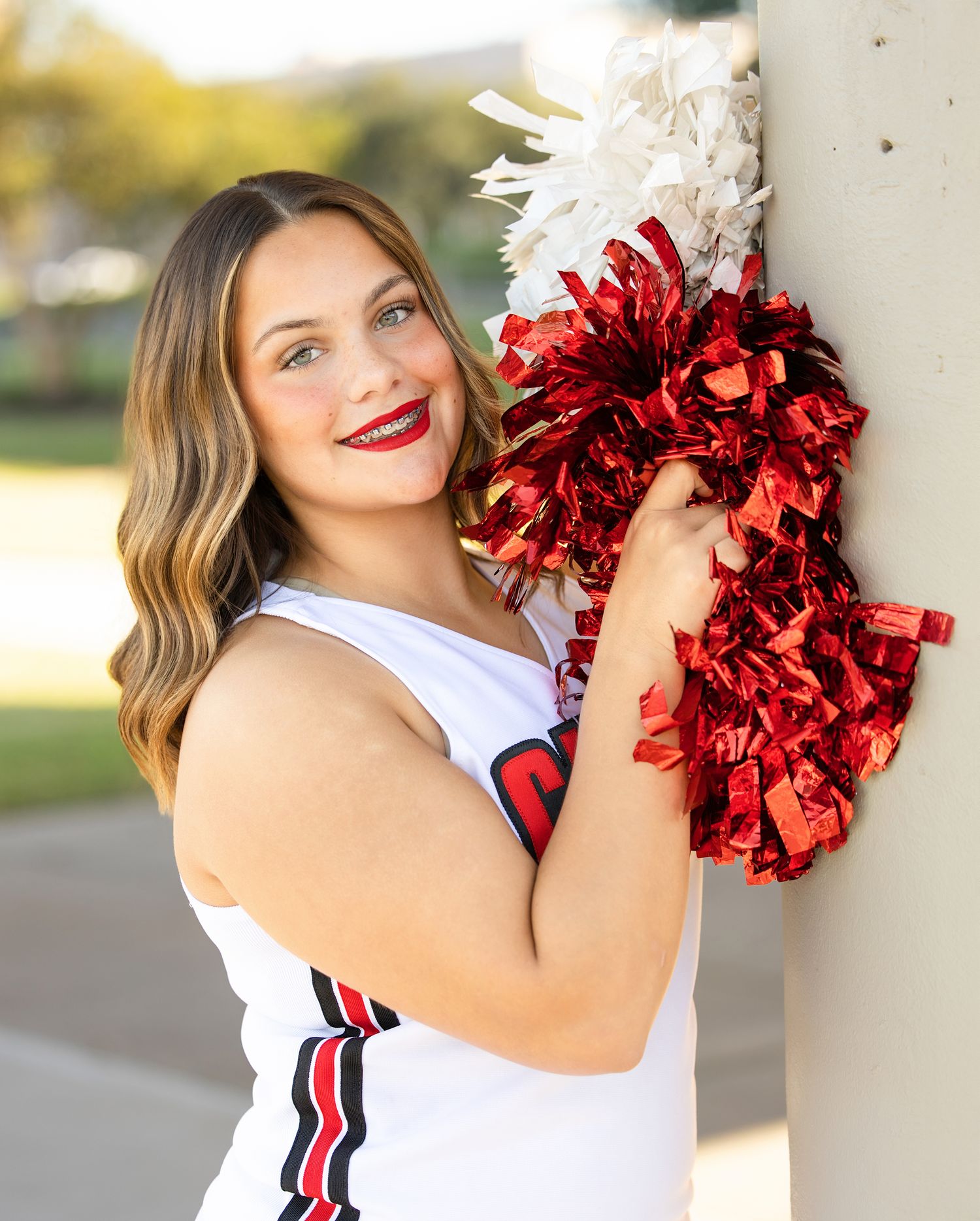 Cheerleader holds red and white pom poms while posing near white columns outdoors.