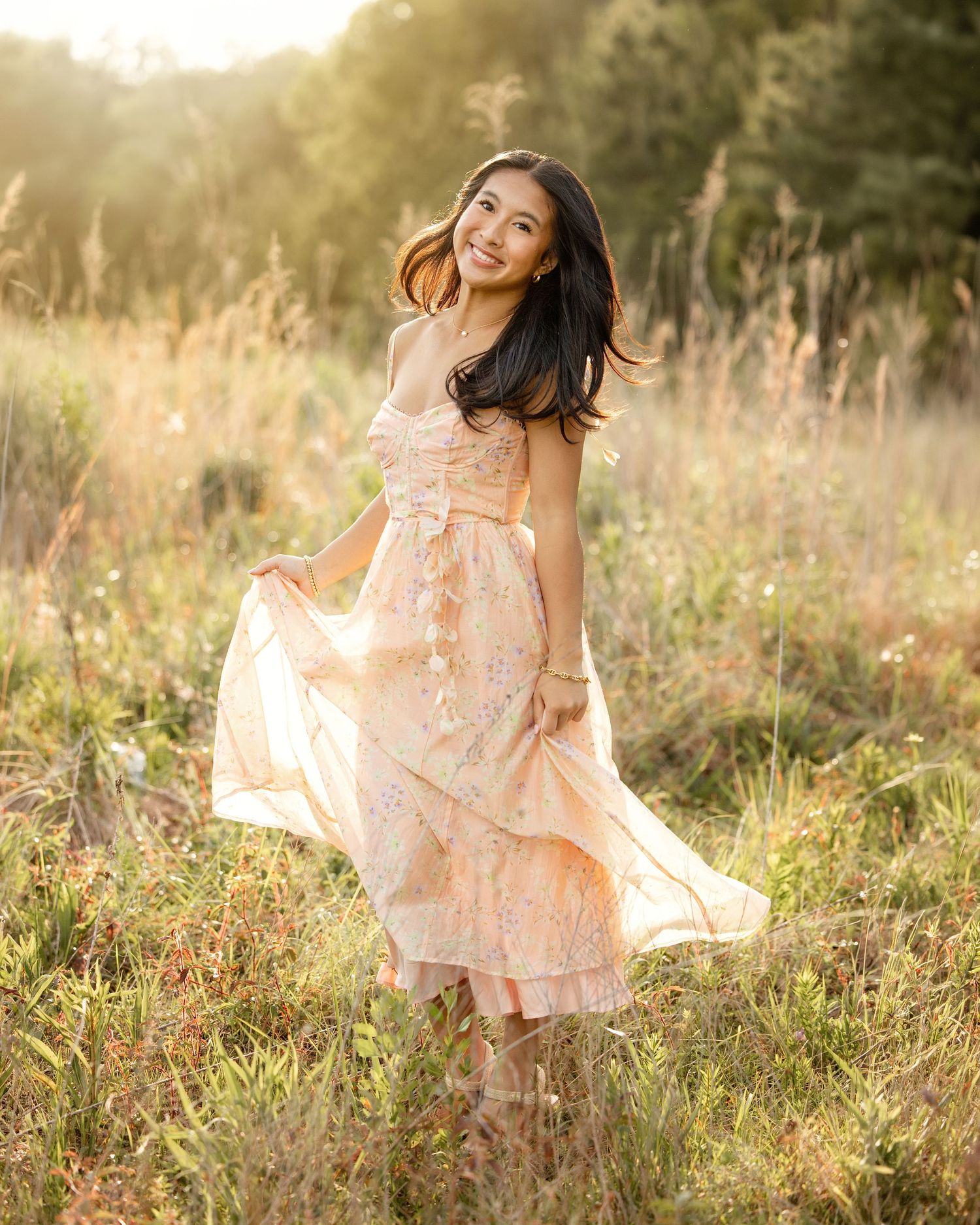 Young woman in a flowy peach floral dress posing in a sunlit meadow with tall golden grasses.