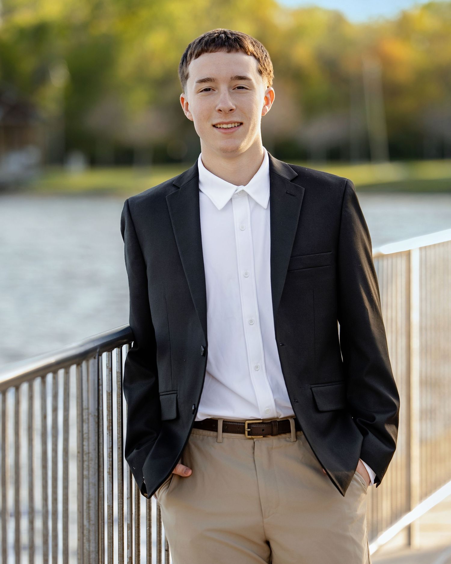 Young man in black blazer and white shirt smiling outdoors near a waterfront railing on a sunny day.