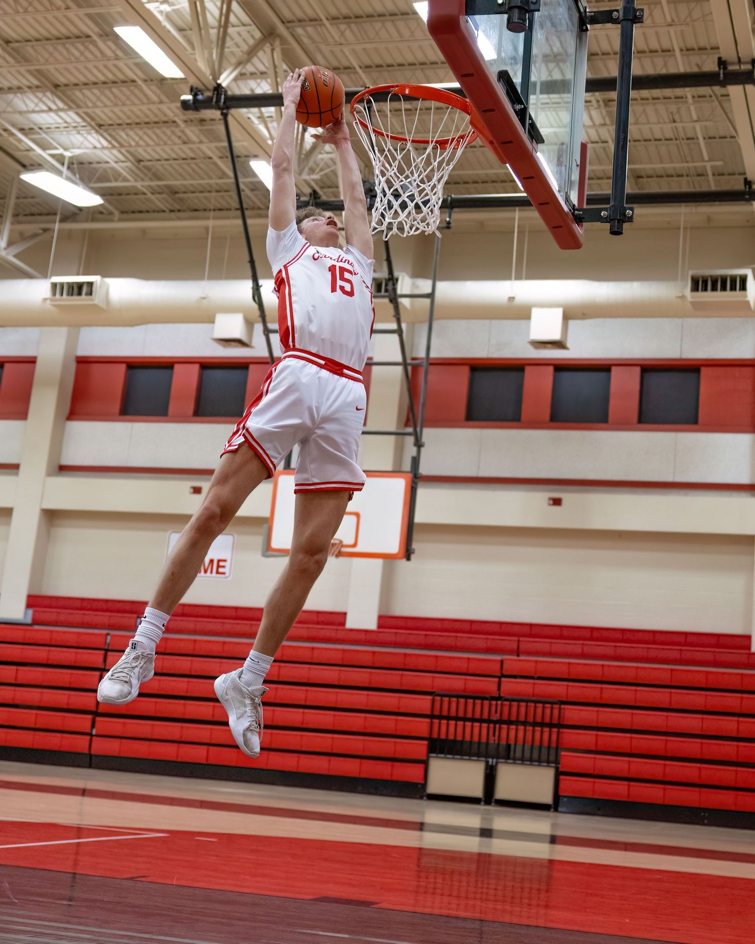 Basketball player in white and red jersey number 15 leaps high for a slam dunk in a school gymnasium.