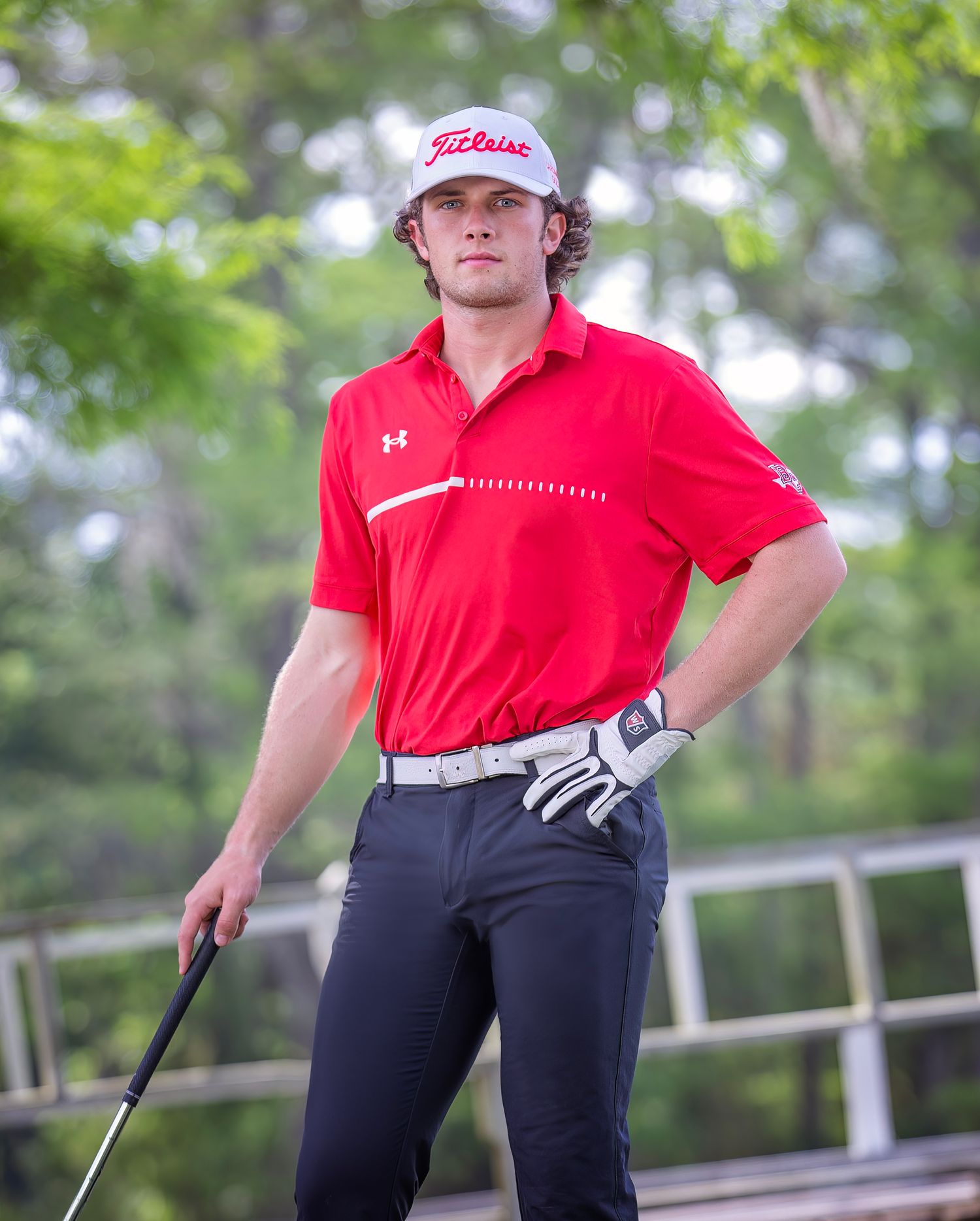 A golfer in red and black attire stands on a golf course with trees in the background.