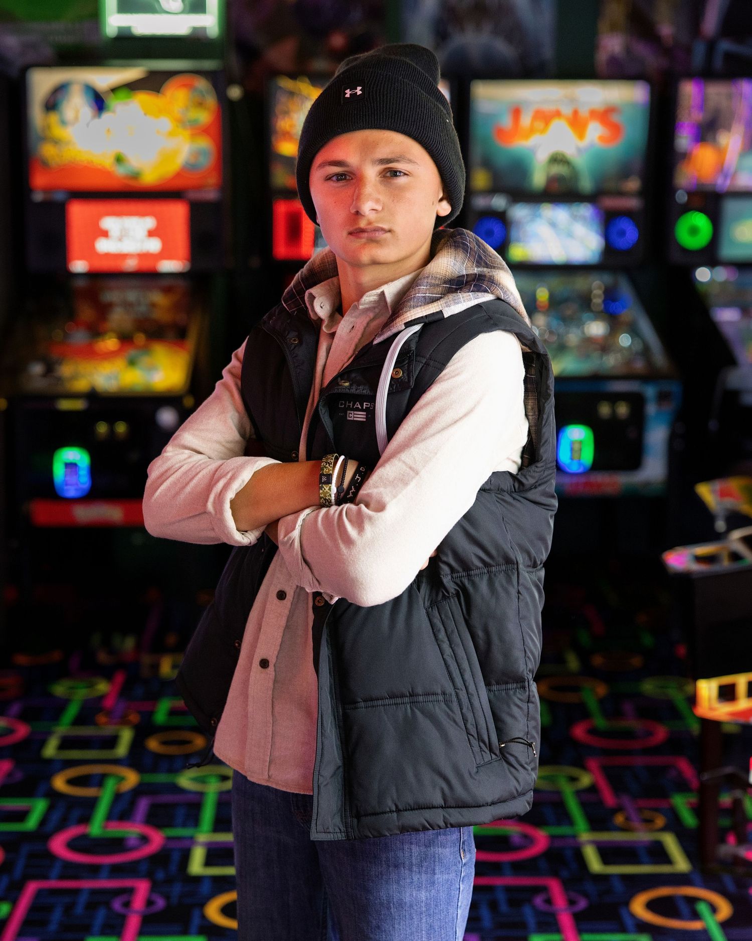 Young man in black vest and beanie stands arms crossed in a colorful retro arcade game room.