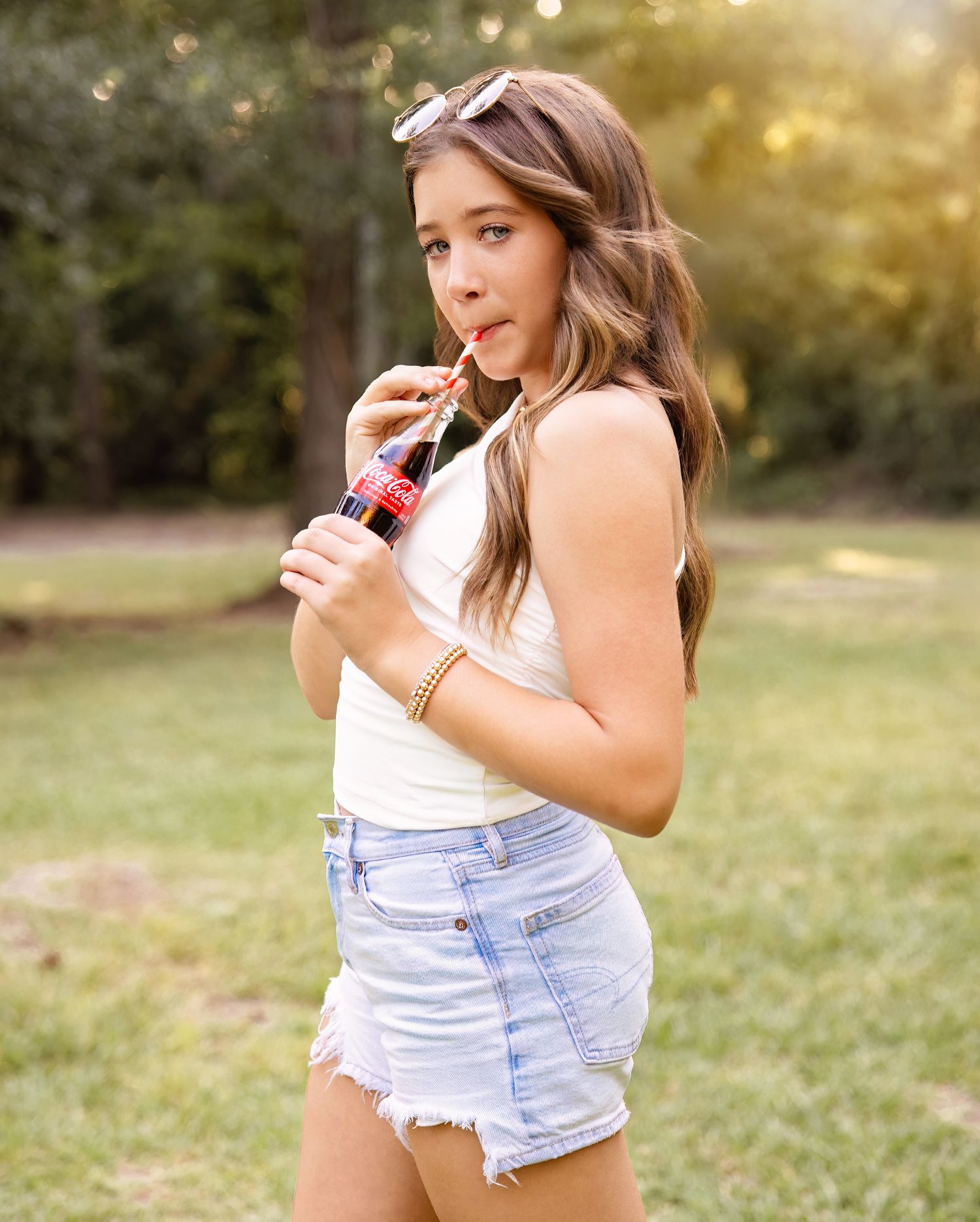A person in denim shorts and white tank top poses casually in a sunlit outdoor setting with bokeh effects.