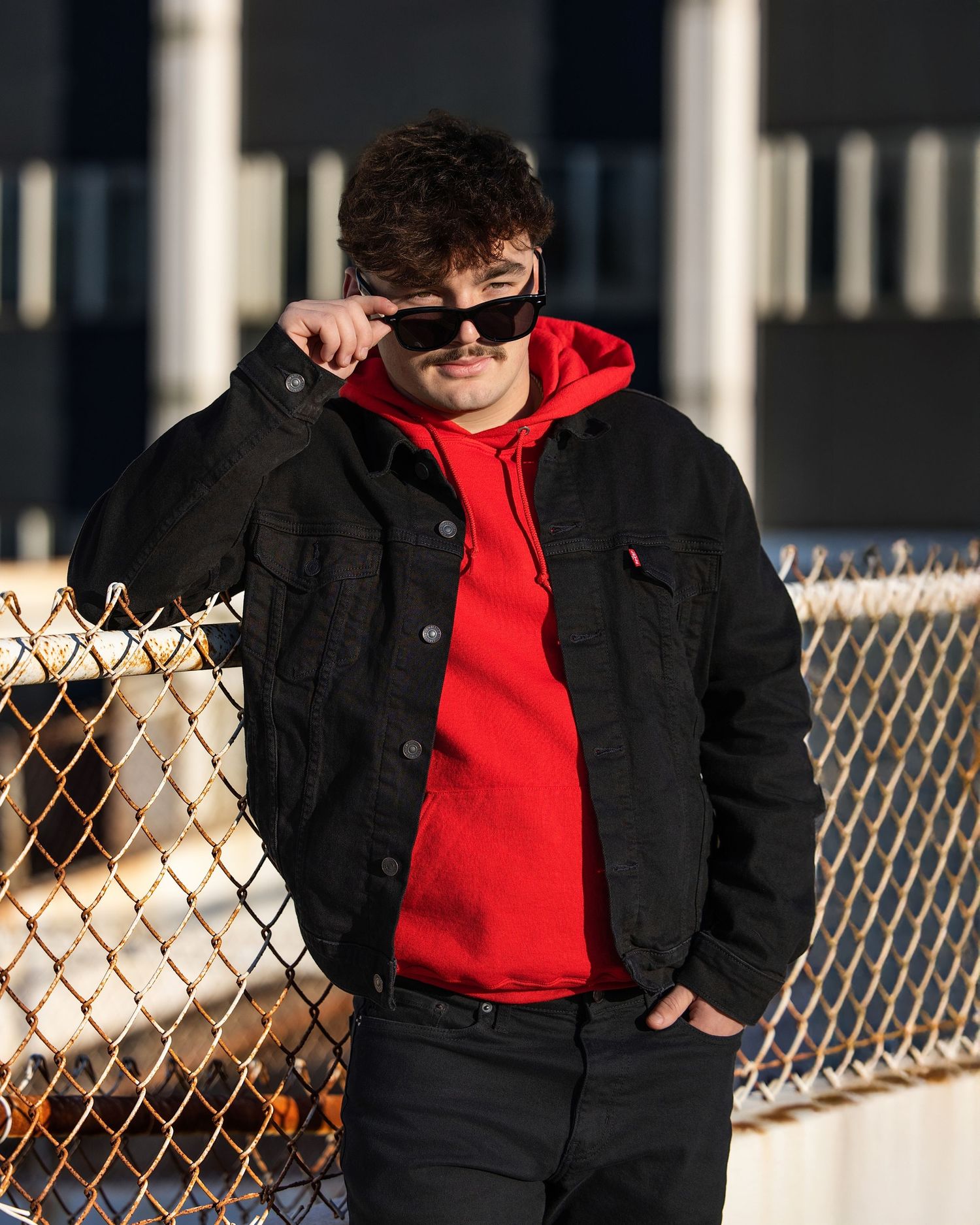 Person wearing a black jacket and red hoodie leans against a chain link fence in an outdoor setting.