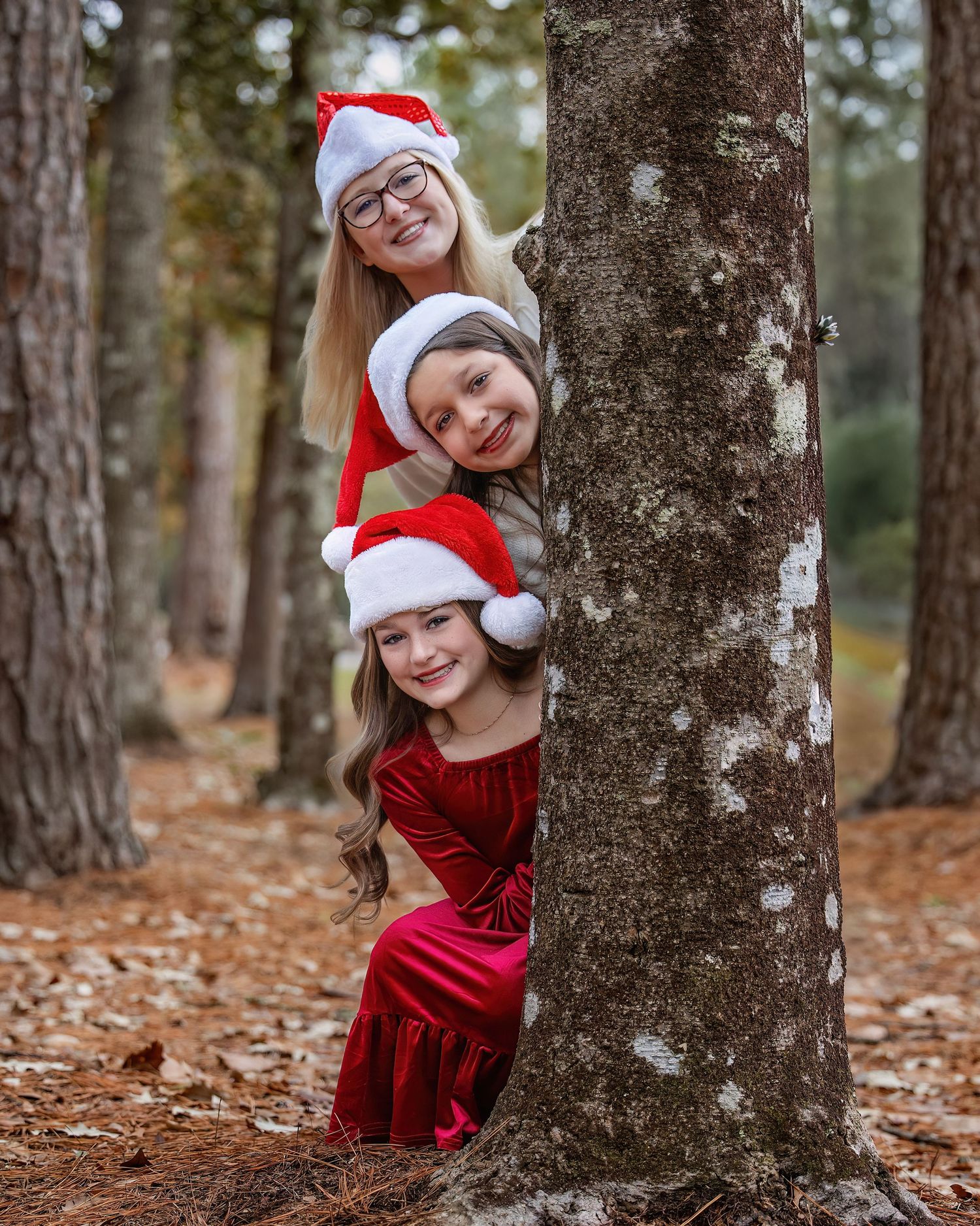 Three people wearing Santa hats peek playfully from behind a tree trunk in a forest covered with fallen autumn leaves.