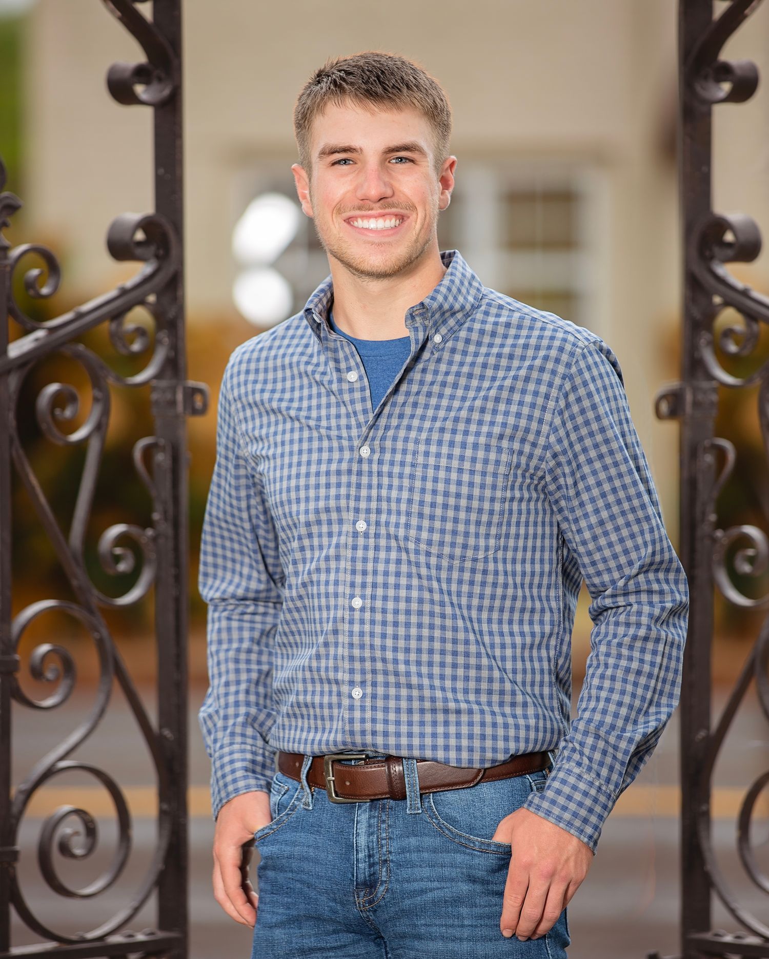 Person wearing a blue checkered button-down shirt and jeans poses by decorative iron railings.