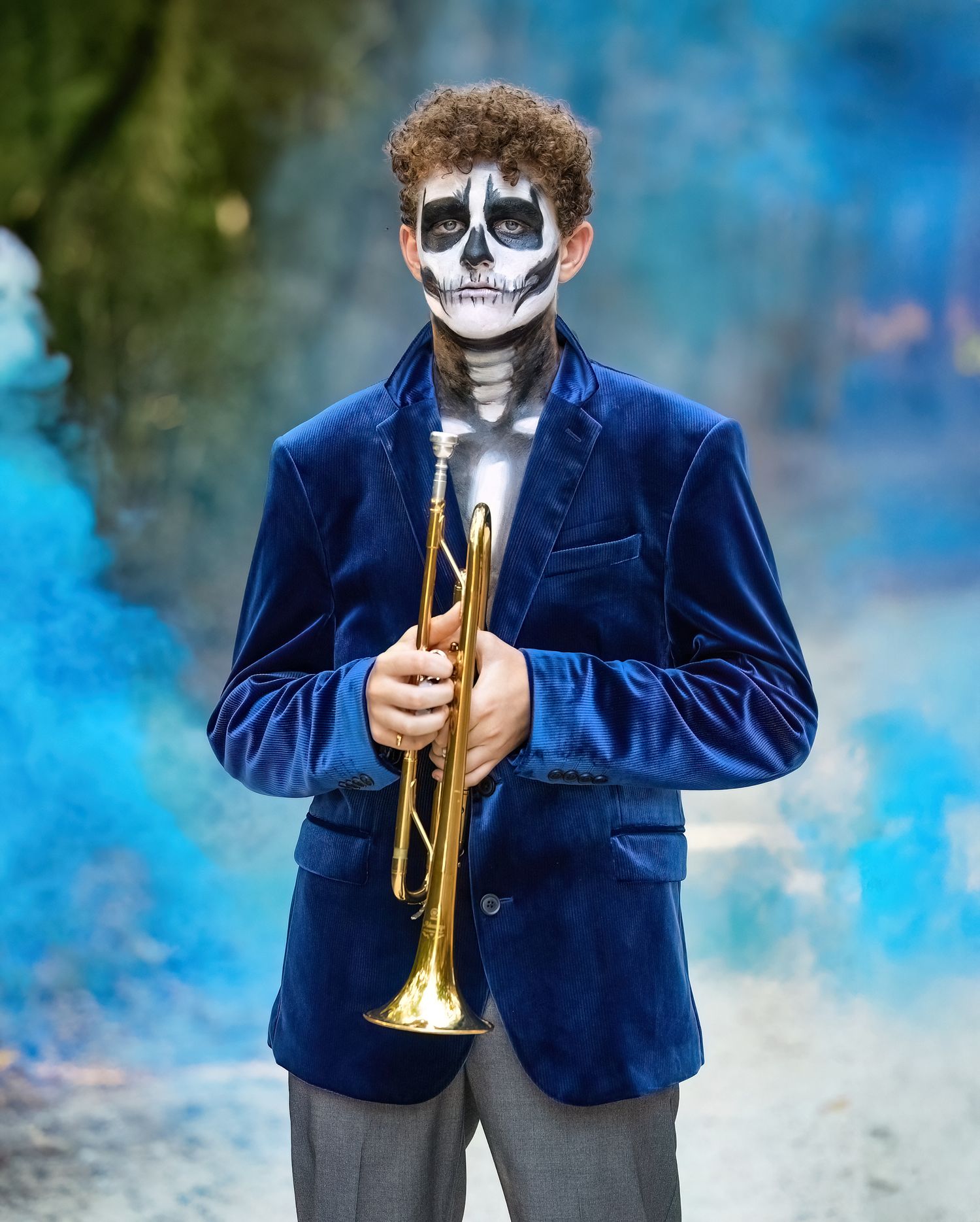 Musician in blue velvet jacket and skeleton face paint plays trumpet with blue smoke effects in background.