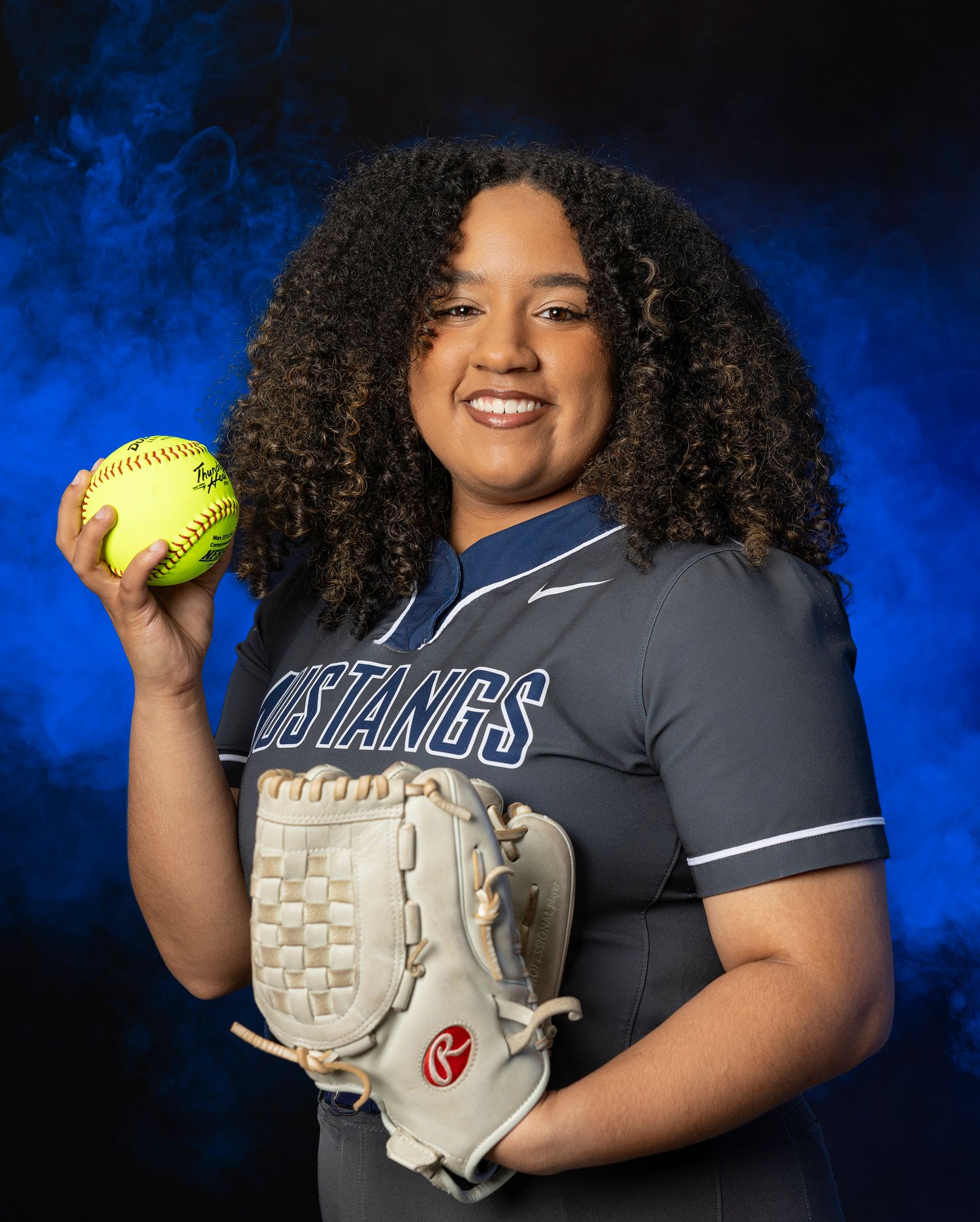 Softball player in gray uniform holds up yellow ball and white glove against dramatic blue backdrop.