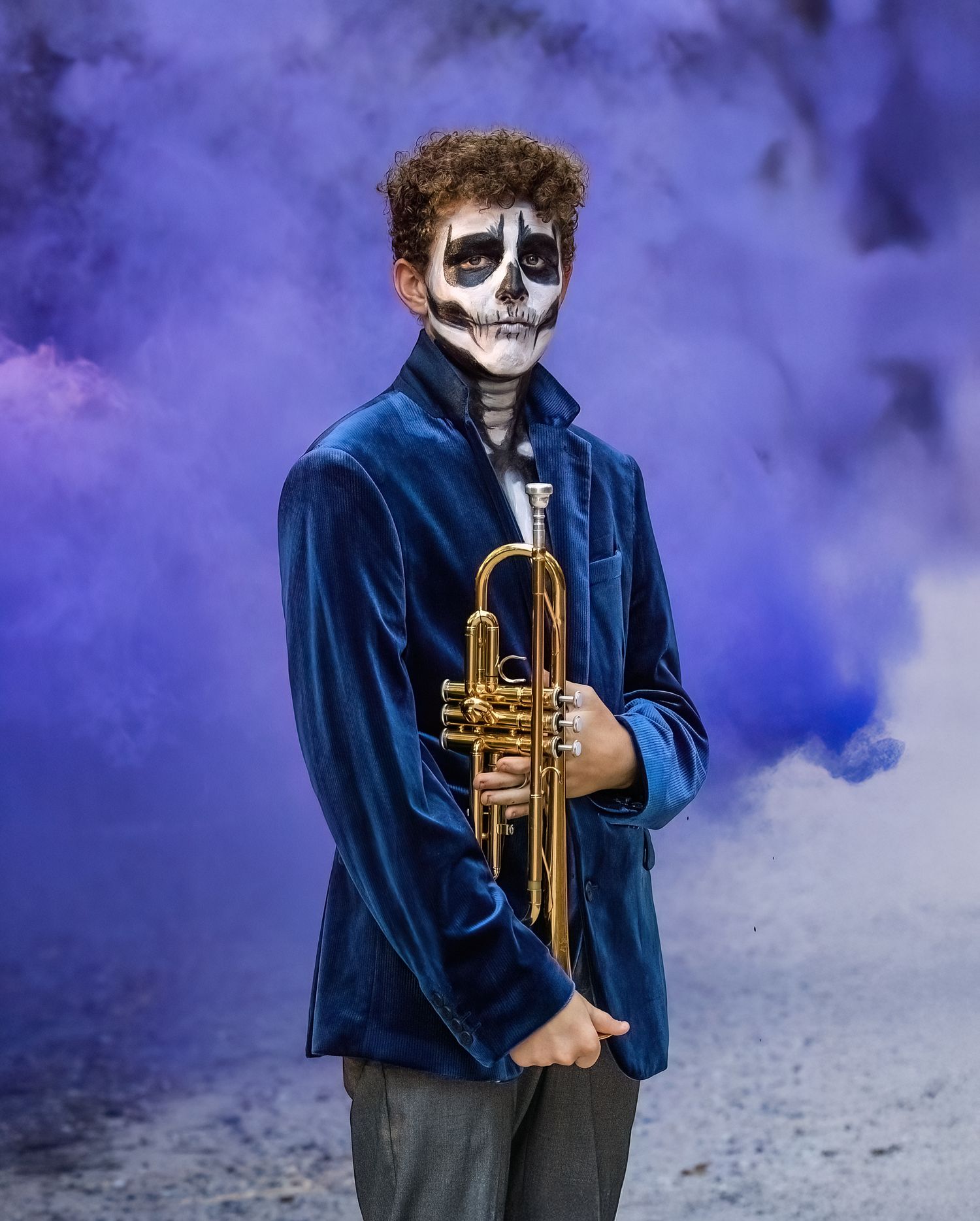 A figure in blue velvet holds a trumpet amid purple smoke effects with skeleton face paint.