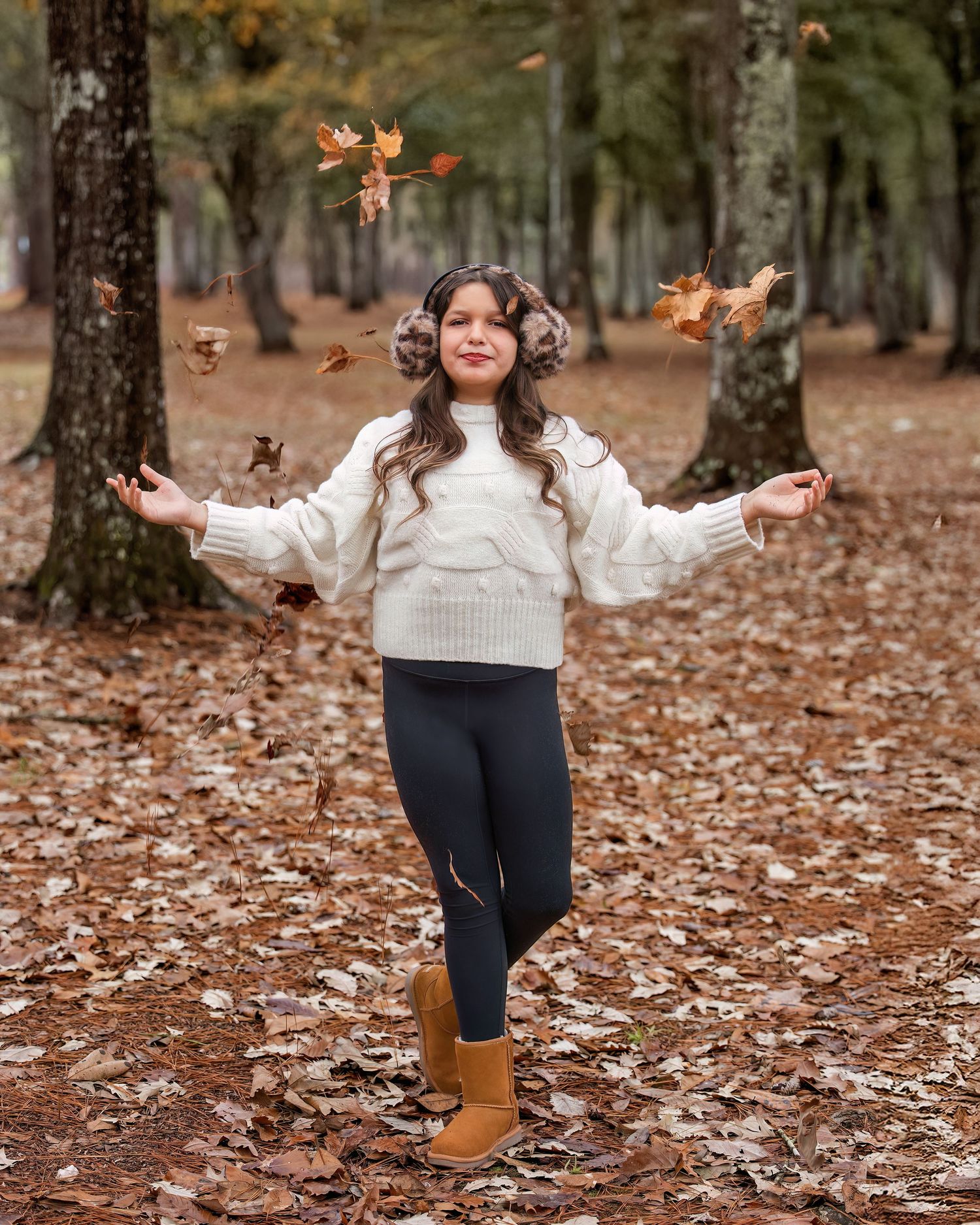 Person in white sweater playfully tosses autumn leaves in the air while standing in a forest.