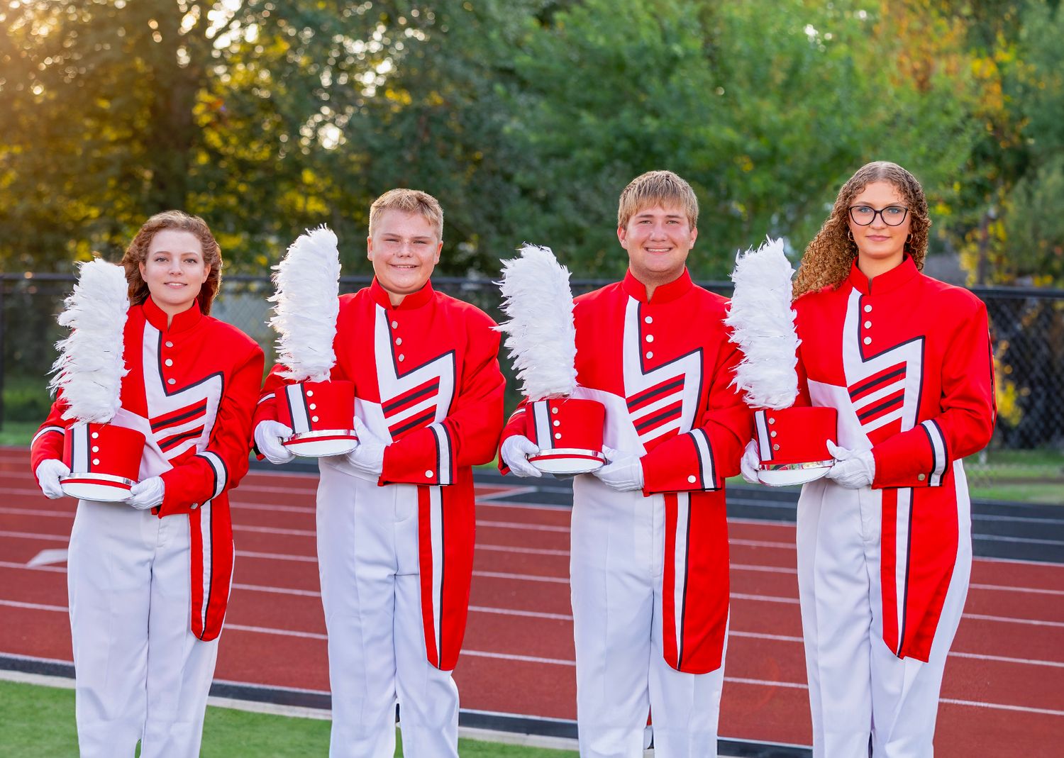 High school marching band members in red and white uniforms stand in formation on an outdoor track field.