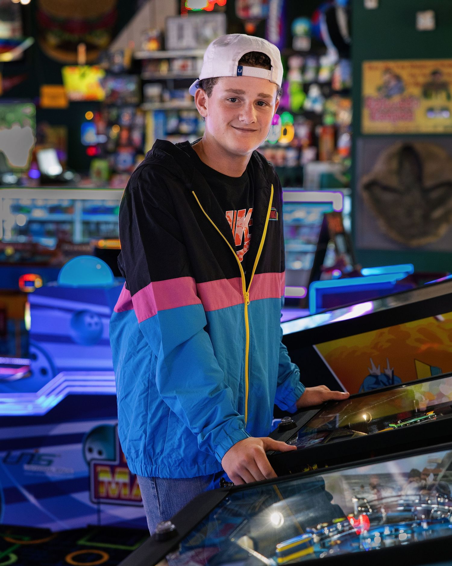 Teen boy in retro windbreaker and backwards cap smiles while playing pinball at a colorful arcade.