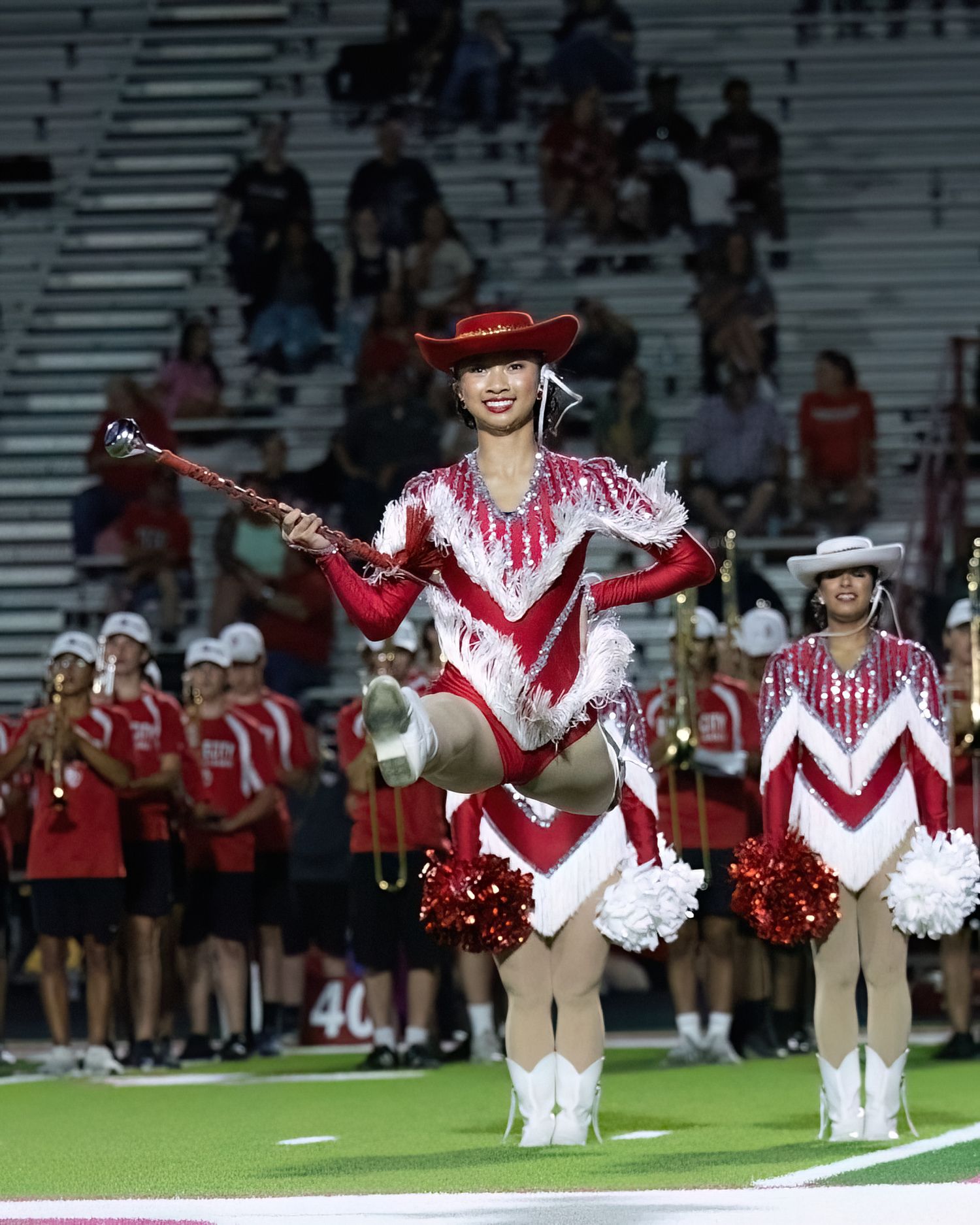 Cheerleaders in red and white uniforms perform synchronized high kicks during a football game halftime show.
