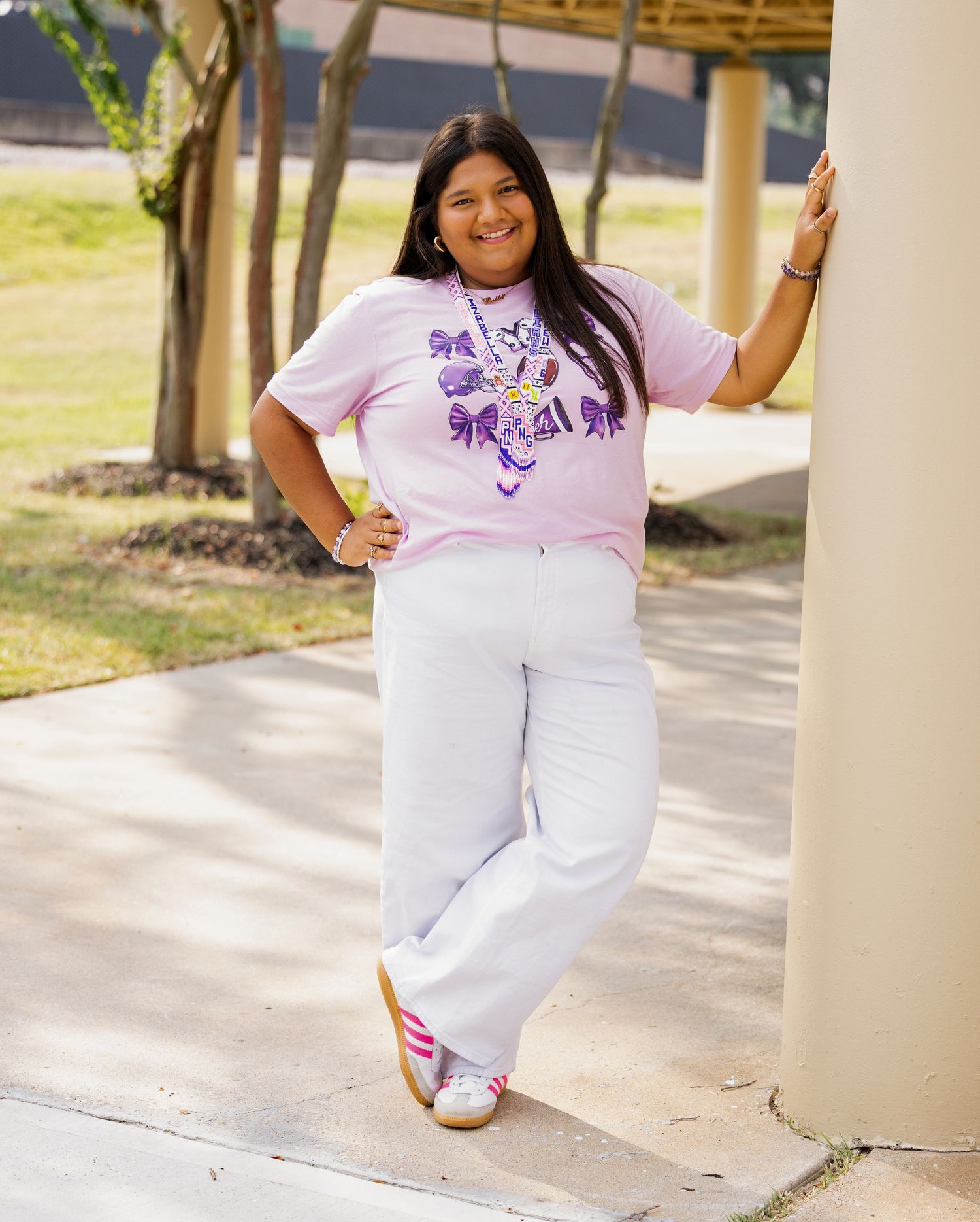 A person in a white and purple t-shirt poses cheerfully in front of a covered walkway on a sunny day.