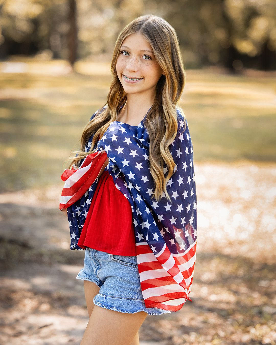 Young girl wearing an American flag print top with denim shorts, smiling outdoors in a park setting.