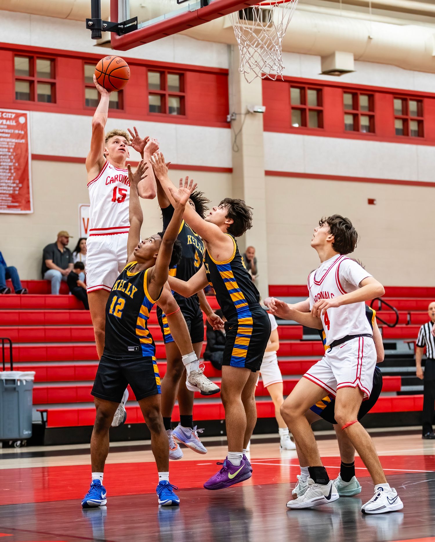 High school basketball players compete for a rebound during an intense game in a gymnasium with red bleachers.