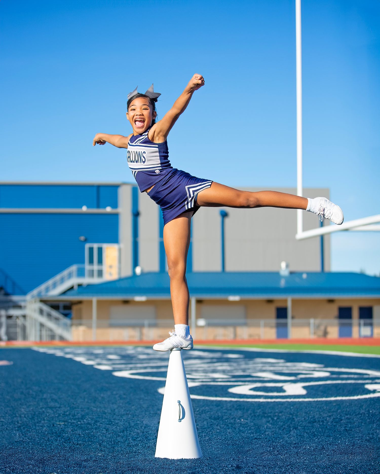 A cheerleader performs a high kick balance pose on a blue turf field with goal posts visible in the background.