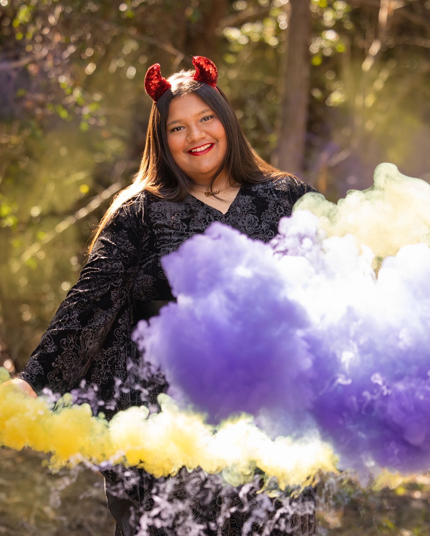 A person in a black dress and red devil horns poses outdoors surrounded by purple and yellow smoke bombs.