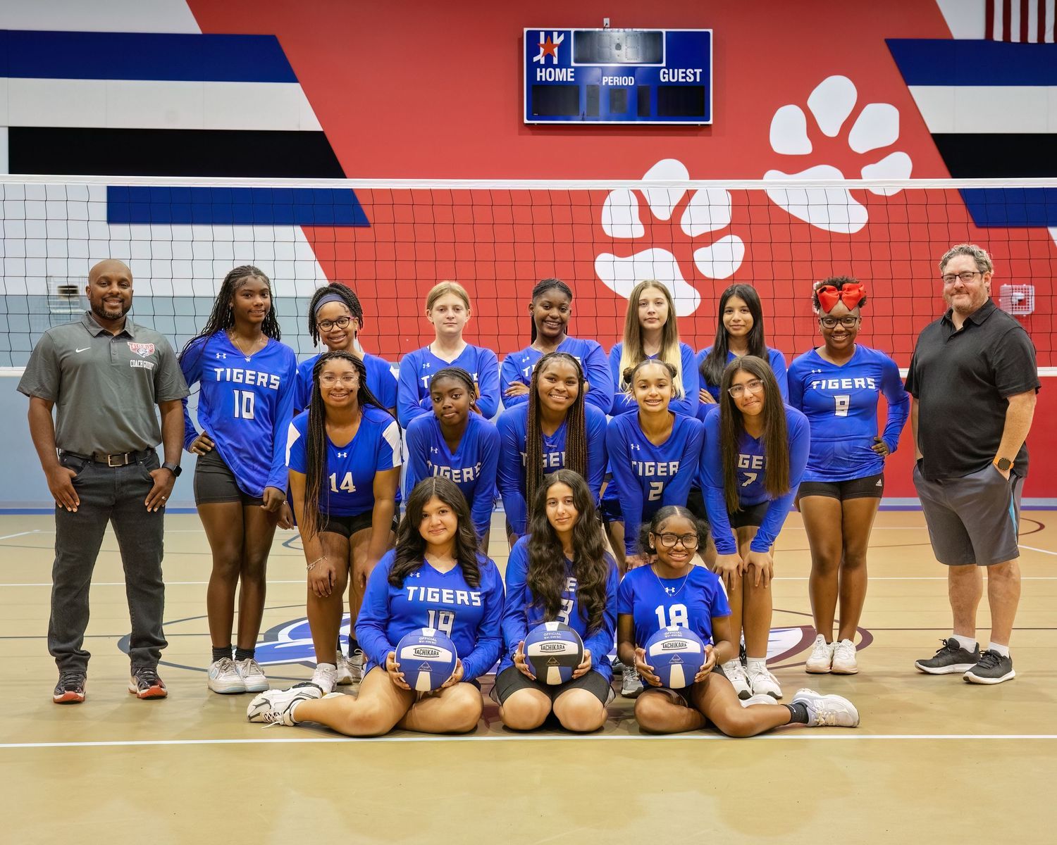 A volleyball team poses for a group photo in blue uniforms on a court with red and black walls.